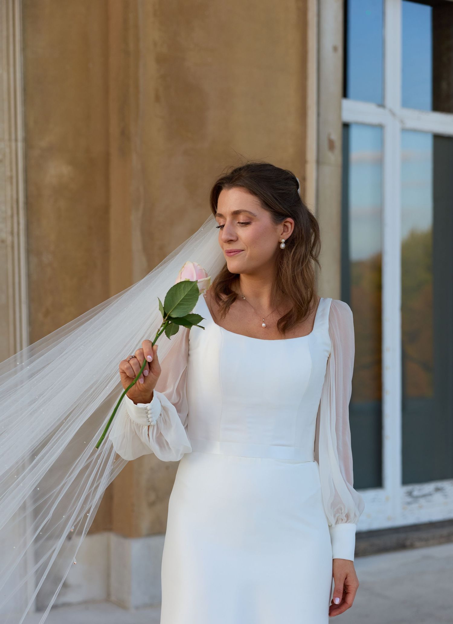A bride in an elegant white gown with sheer sleeves smells a single green stem while posing outdoors.