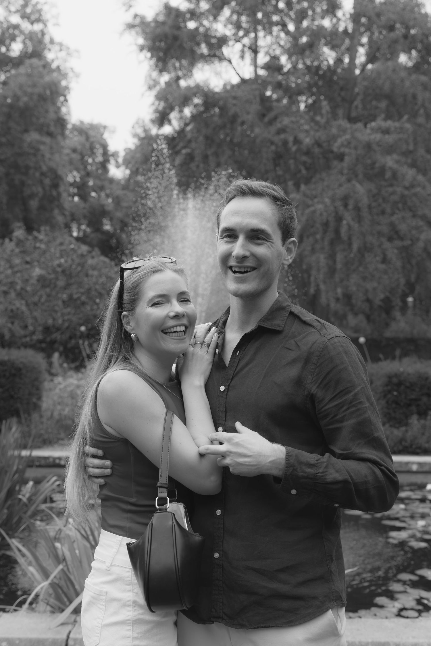 A happy couple embraces and smiles in front of a garden fountain in this black and white portrait photo.