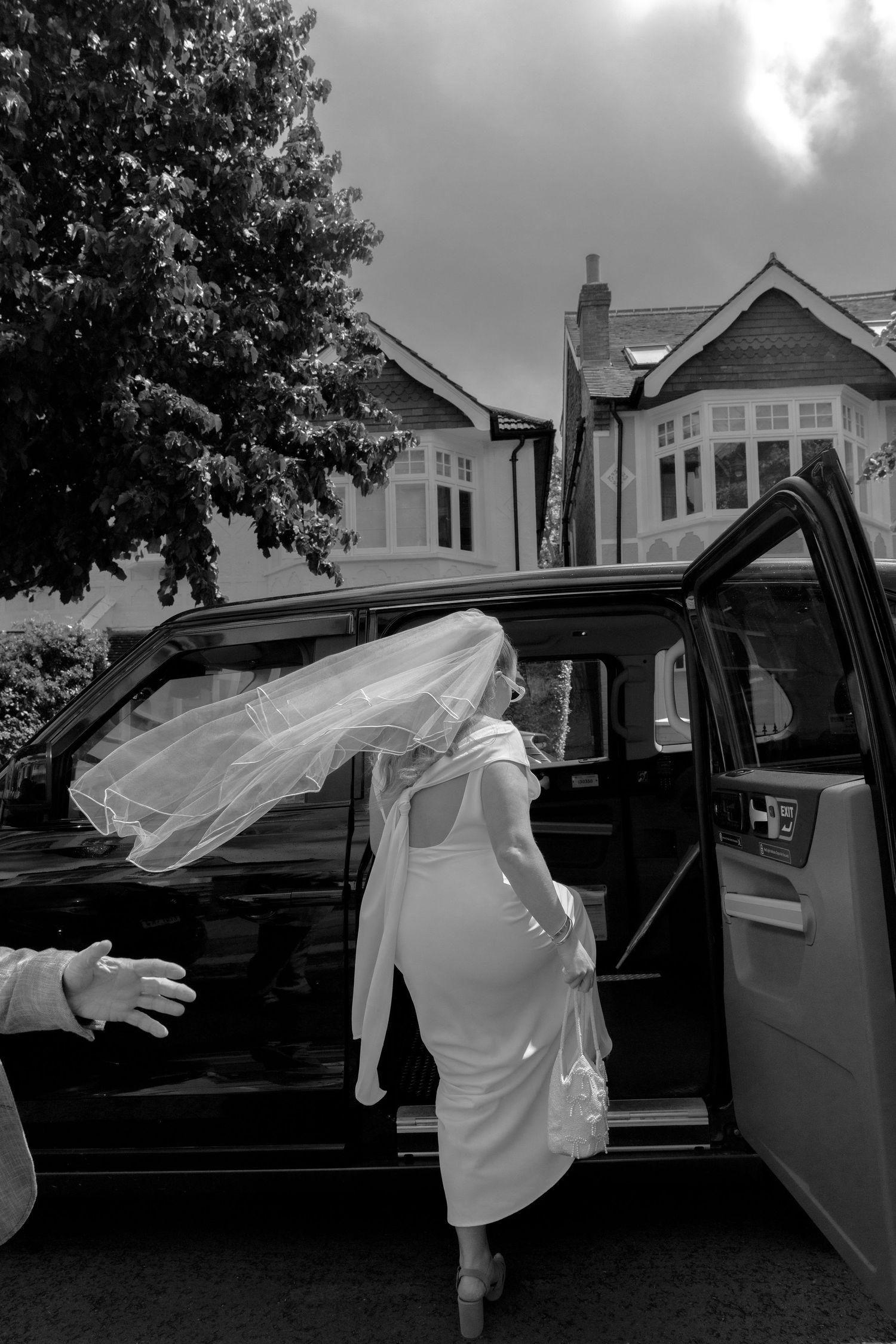 Bride in white dress and veil entering a black car on a windy day, black and white wedding photo.