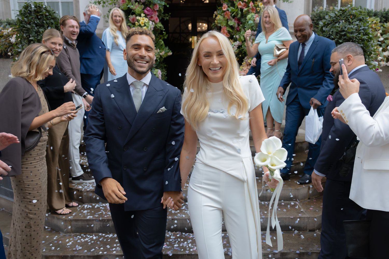 Happy couple walks through confetti shower after wedding ceremony, bride in white jumpsuit holding bouquet, groom in navy suit.
