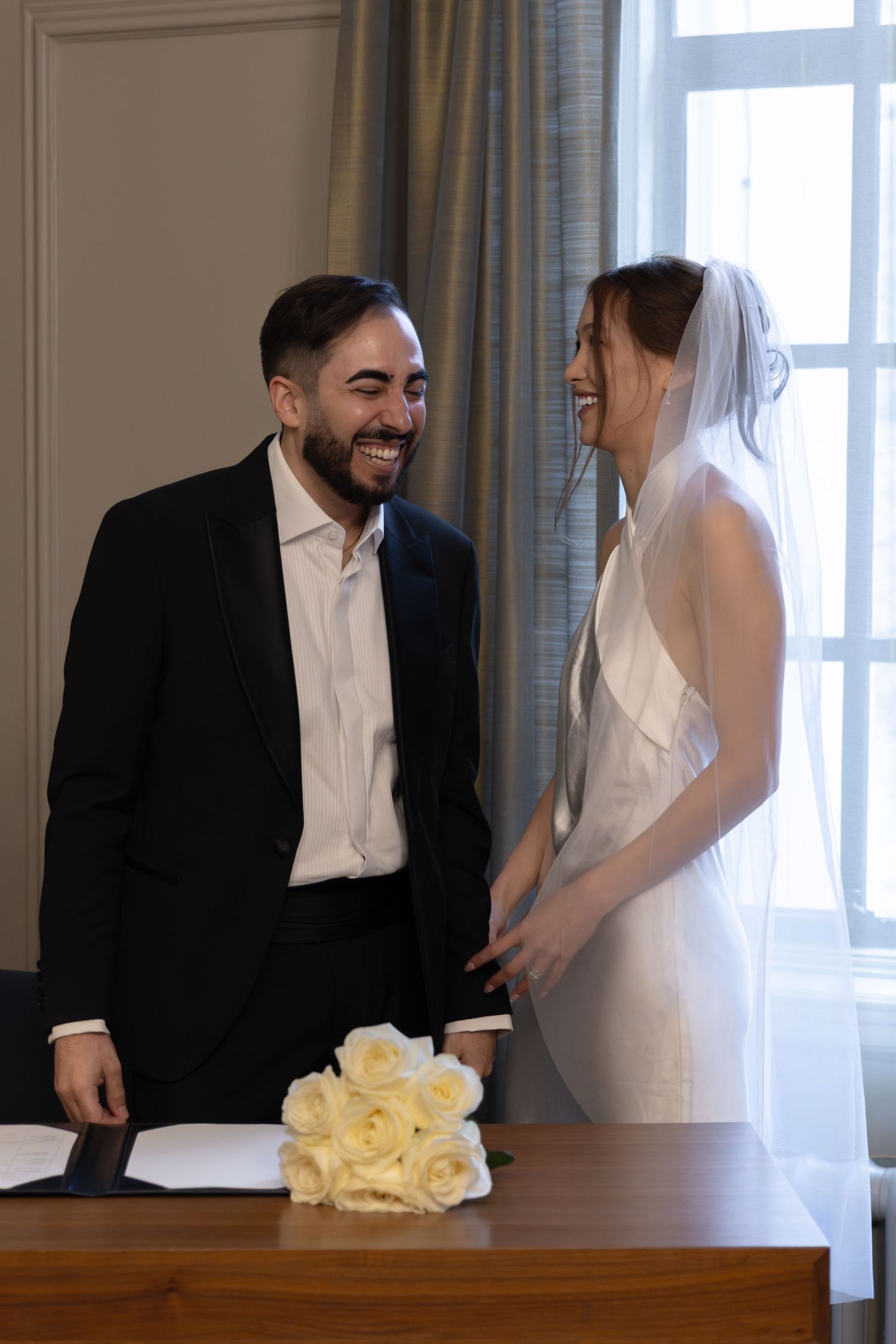 Happy bride and groom laughing together at signing table with white roses bouquet during wedding ceremony.