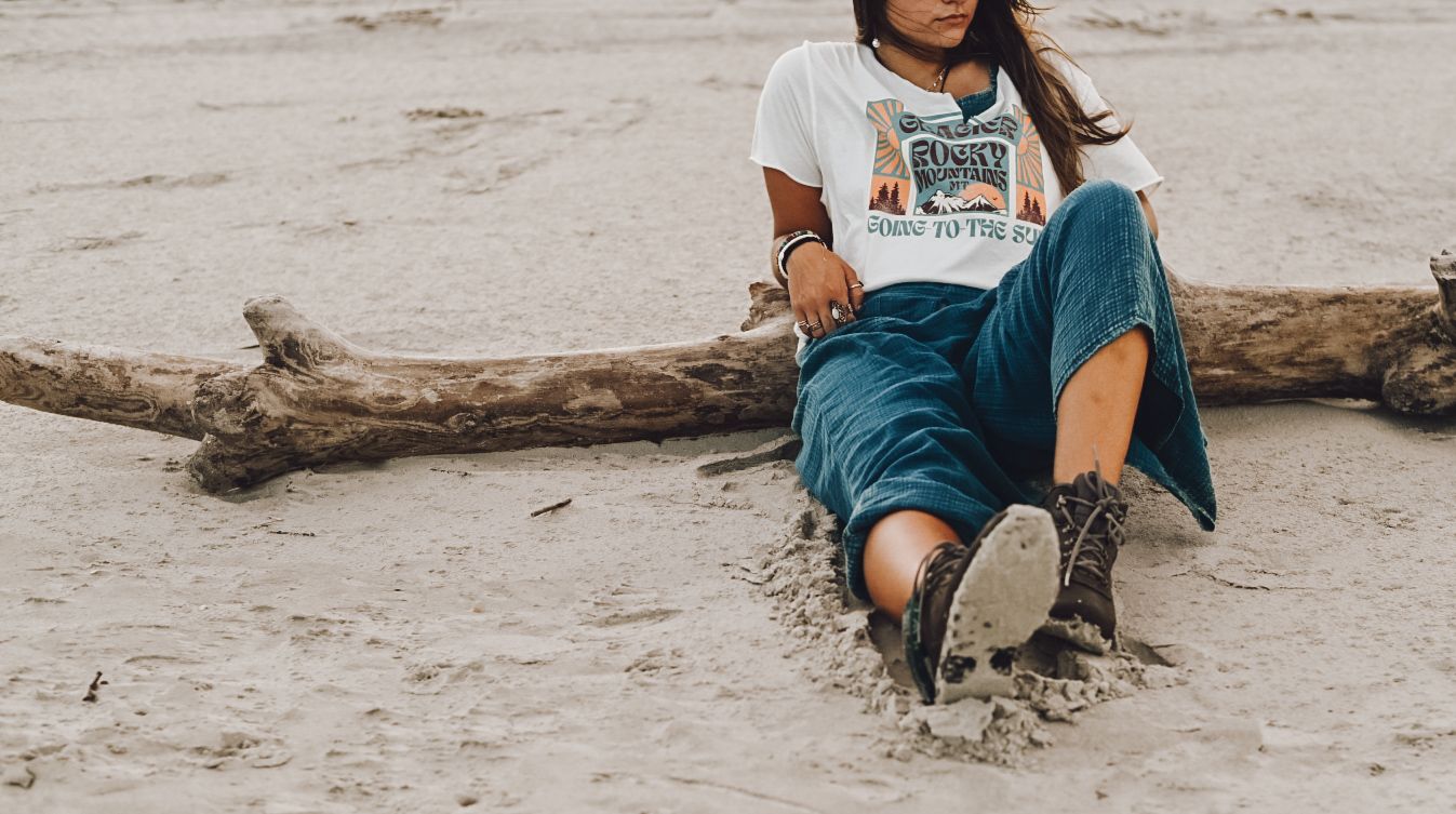Person wearing blue jeans and graphic tee relaxes on sandy beach next to driftwood log.