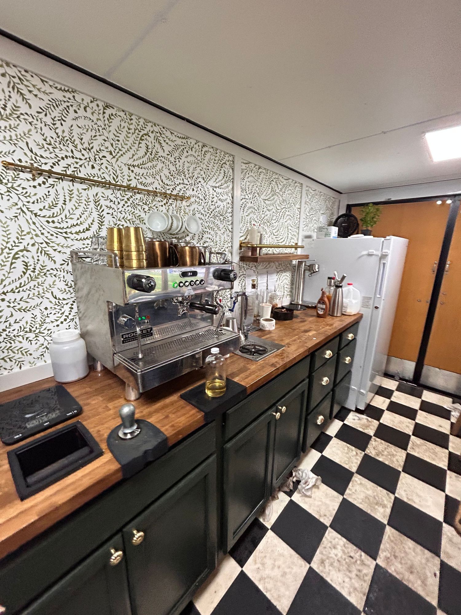 Modern coffee shop interior with black and white checkered floor and wooden countertops against floral wallpaper decor.