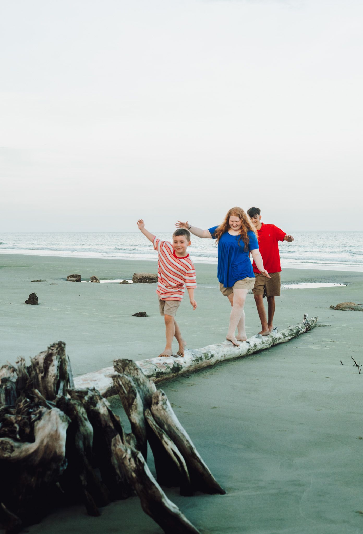 Three people balance and play on a large driftwood log at the beach with waves and rocks in the background.