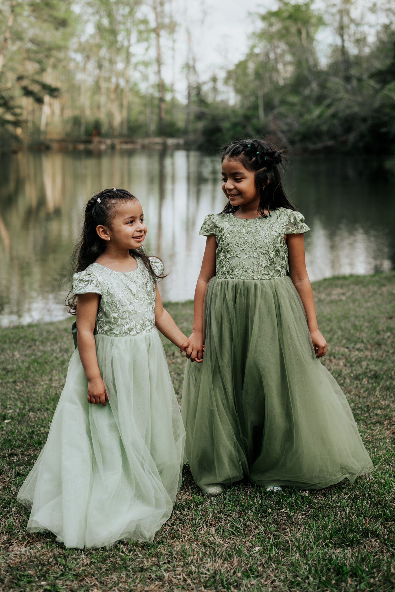 Two young girls in matching sage green lace and tulle gowns hold hands by a tranquil lake in a wooded setting.