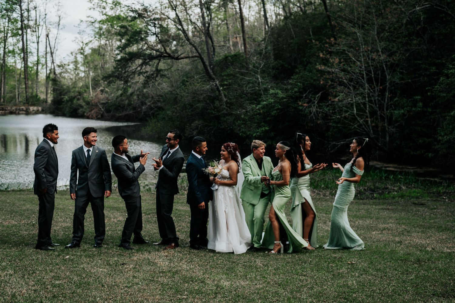 Wedding party in sage green dresses and dark suits pose playfully by a lake in a wooded setting.