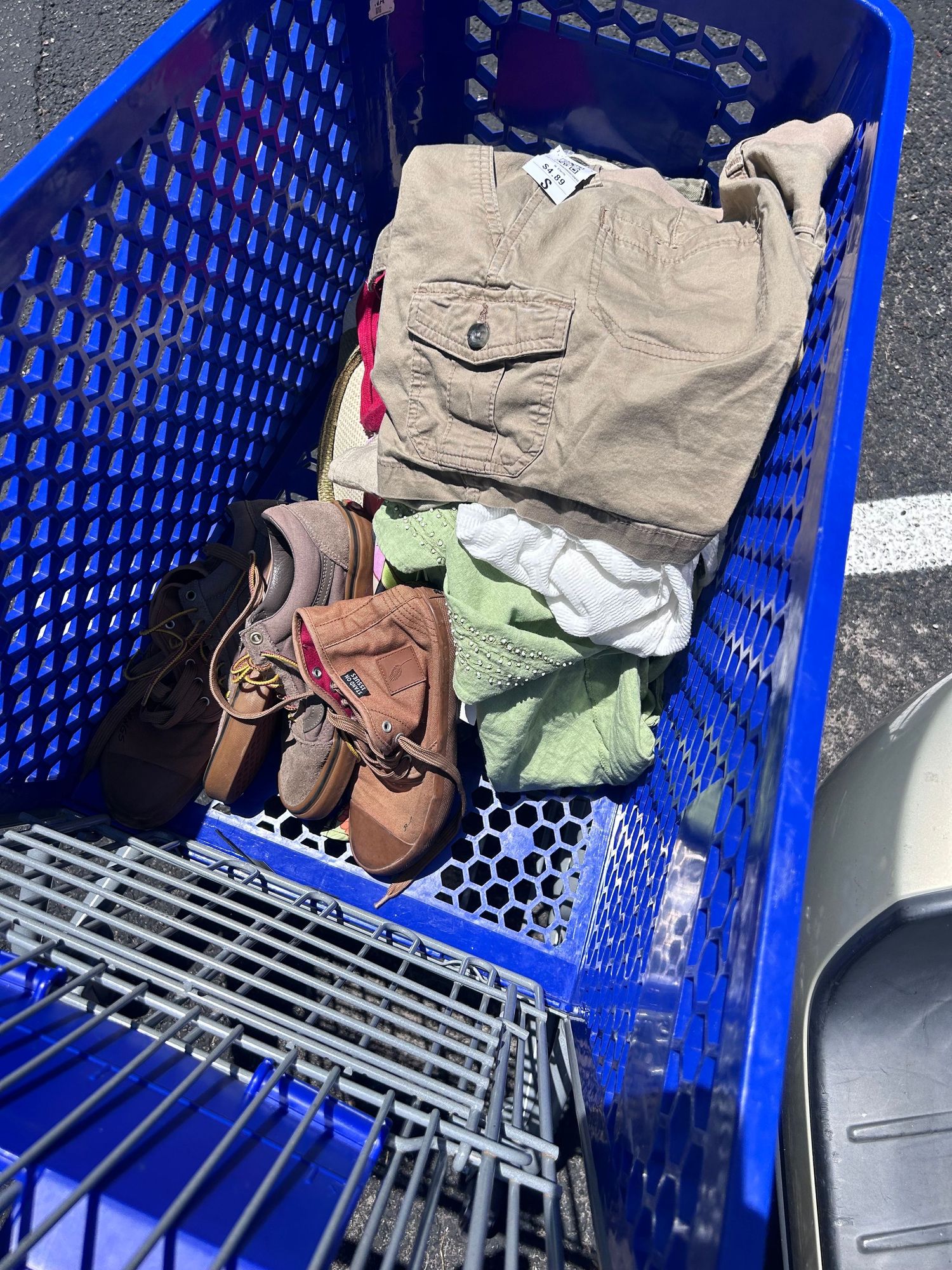 A blue plastic shopping cart containing khaki cargo shorts, brown boots, and folded clothing items.
