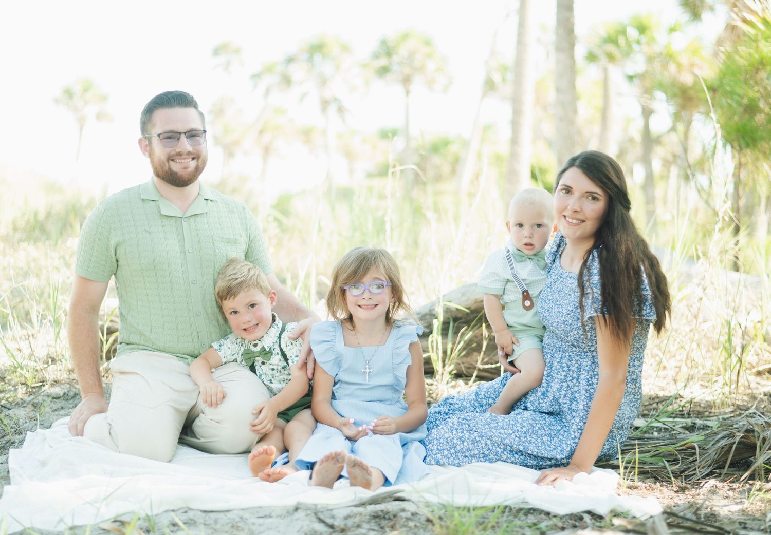 A family wearing pastel colored outfits sits together on a blanket outdoors during a relaxed photo session.