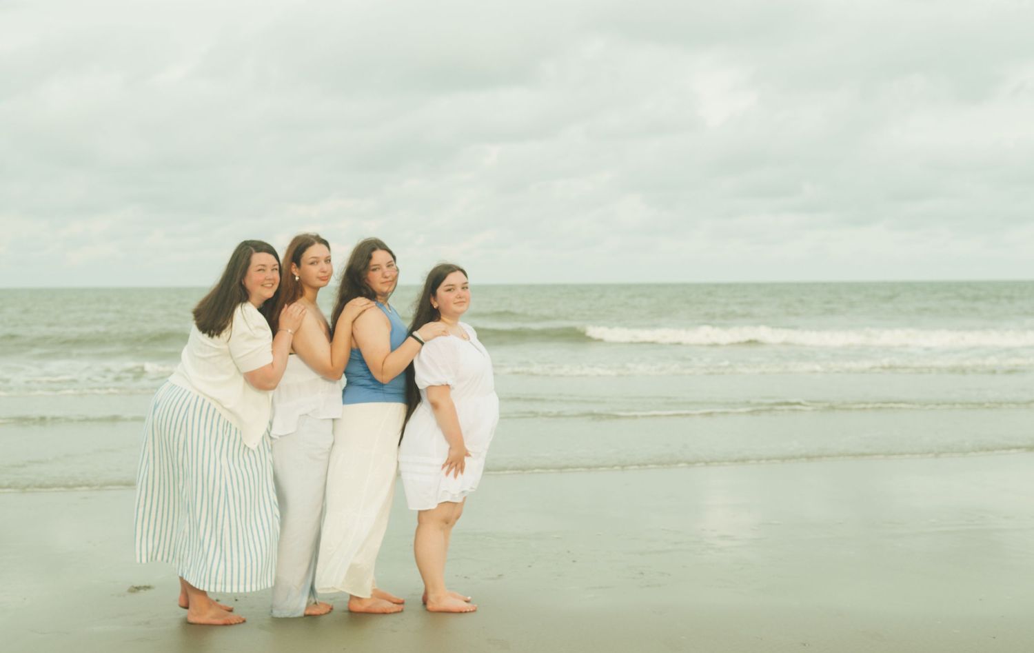 People in light-colored clothing standing together on a cloudy beach with ocean waves and overcast sky in view.