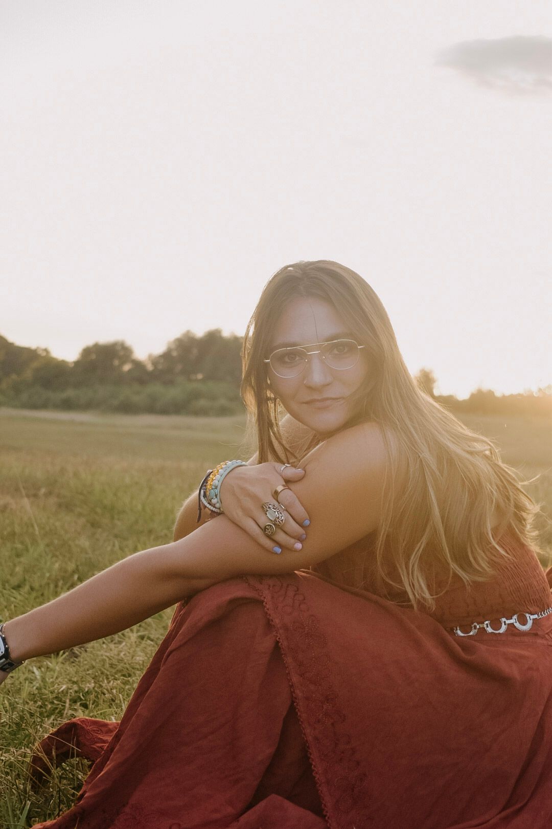 Person in glasses sitting in grassy field at sunset wearing a red dress and jewelry with golden sunlight.