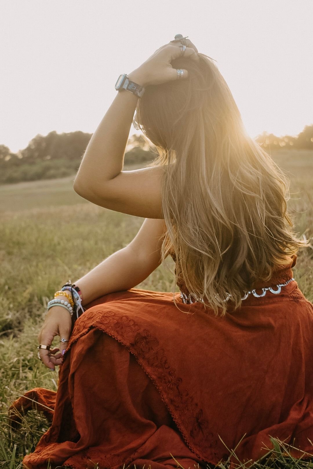 A person in a rust-colored top sits in a grassy field at golden hour, photographed from behind.