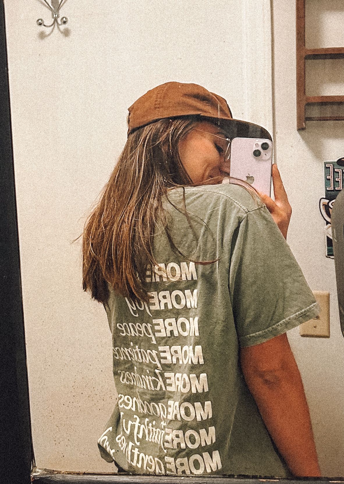 Mirror selfie showing a person in a brown cap and olive green t-shirt with repeating text pattern on the back.