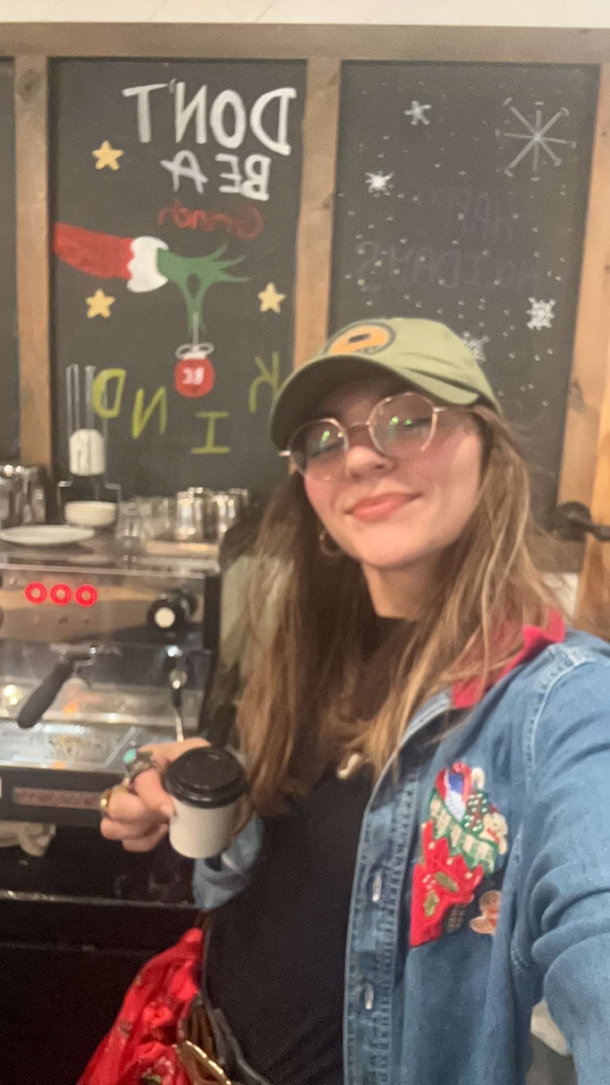 Person in denim jacket and glasses poses with espresso machine at cafe counter with chalkboard wall decorations.