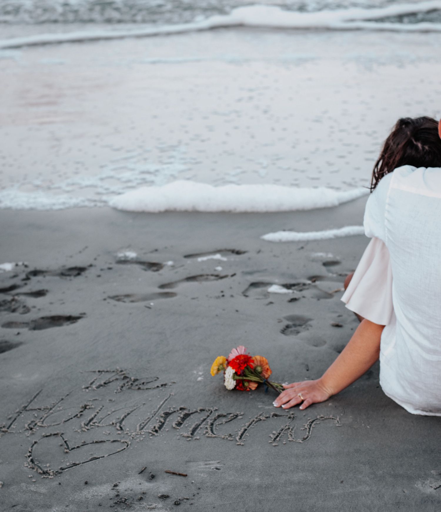 Someone in white sits on a dark sandy beach writing words in the sand beside a small bouquet of colorful flowers.