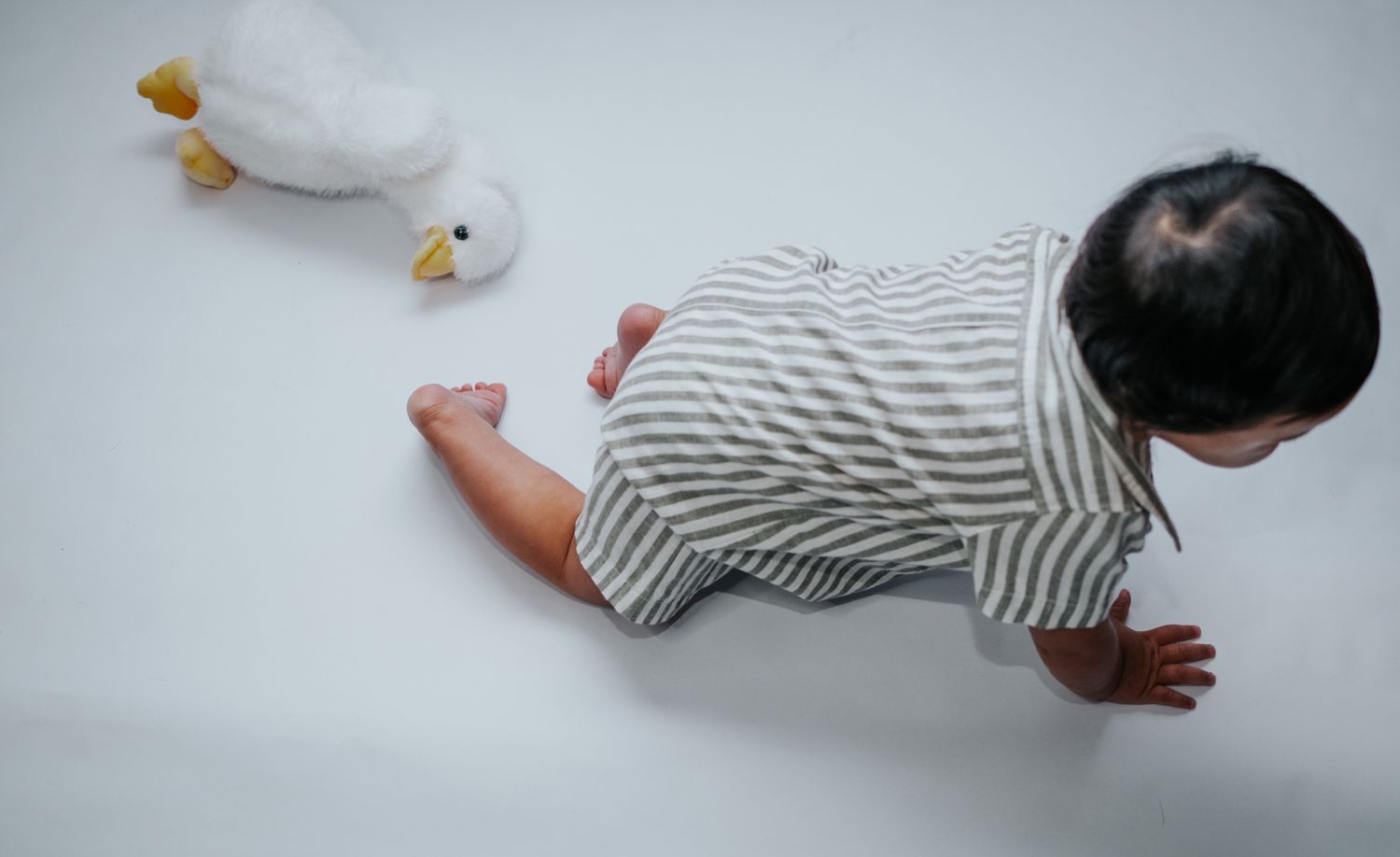 Baby in striped onesie crawling on white surface with toy ducks nearby.