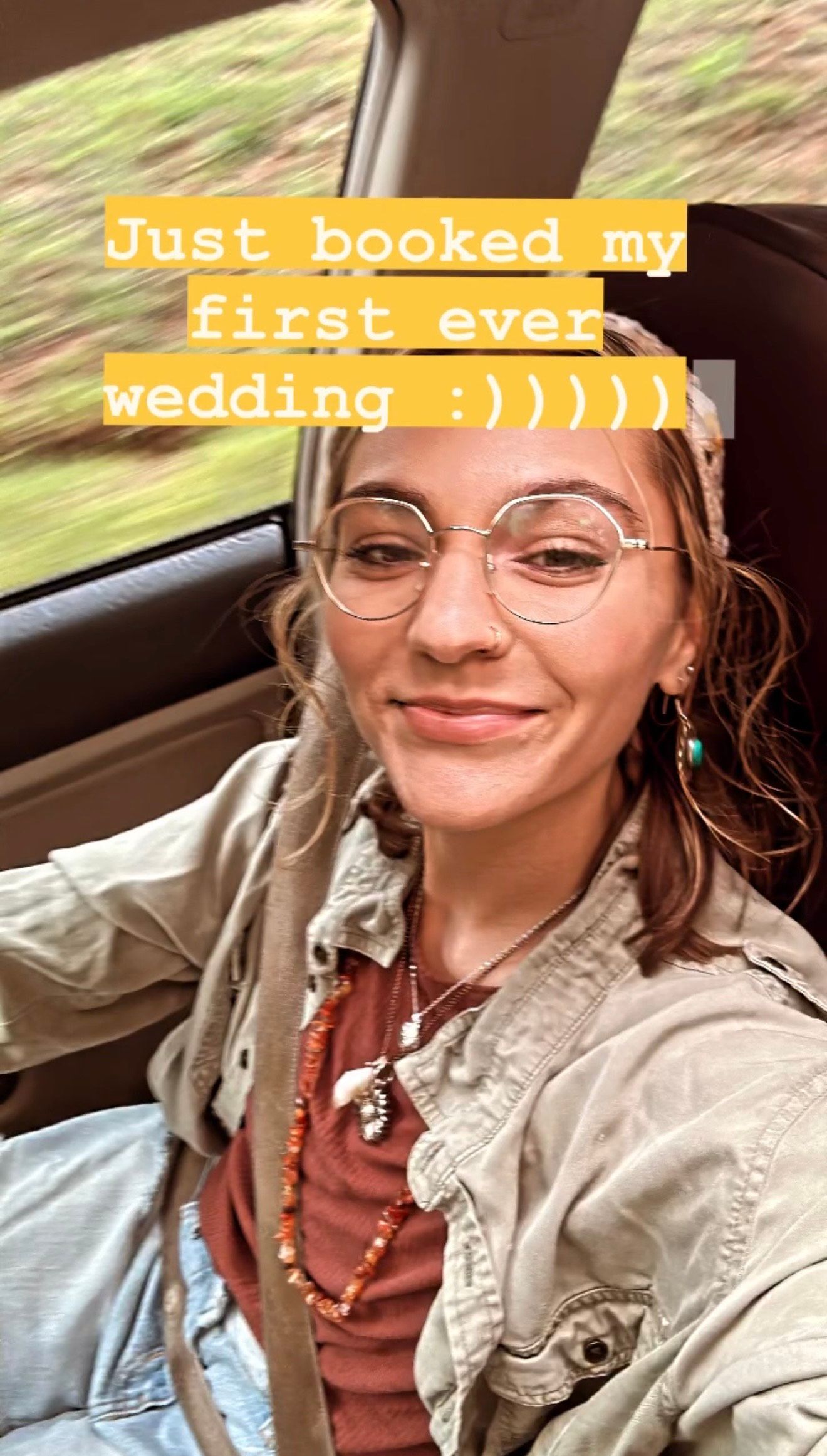 Car selfie shows someone in round glasses and a tan jacket celebrating booking their first wedding photography gig.