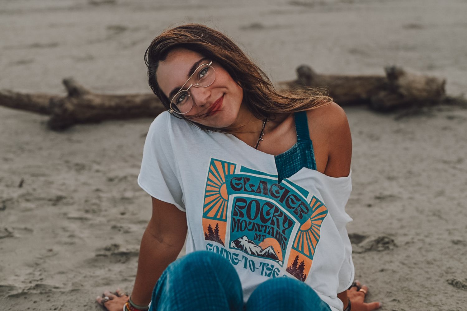 Person in white t-shirt and overalls sits casually on driftwood at sandy beach.