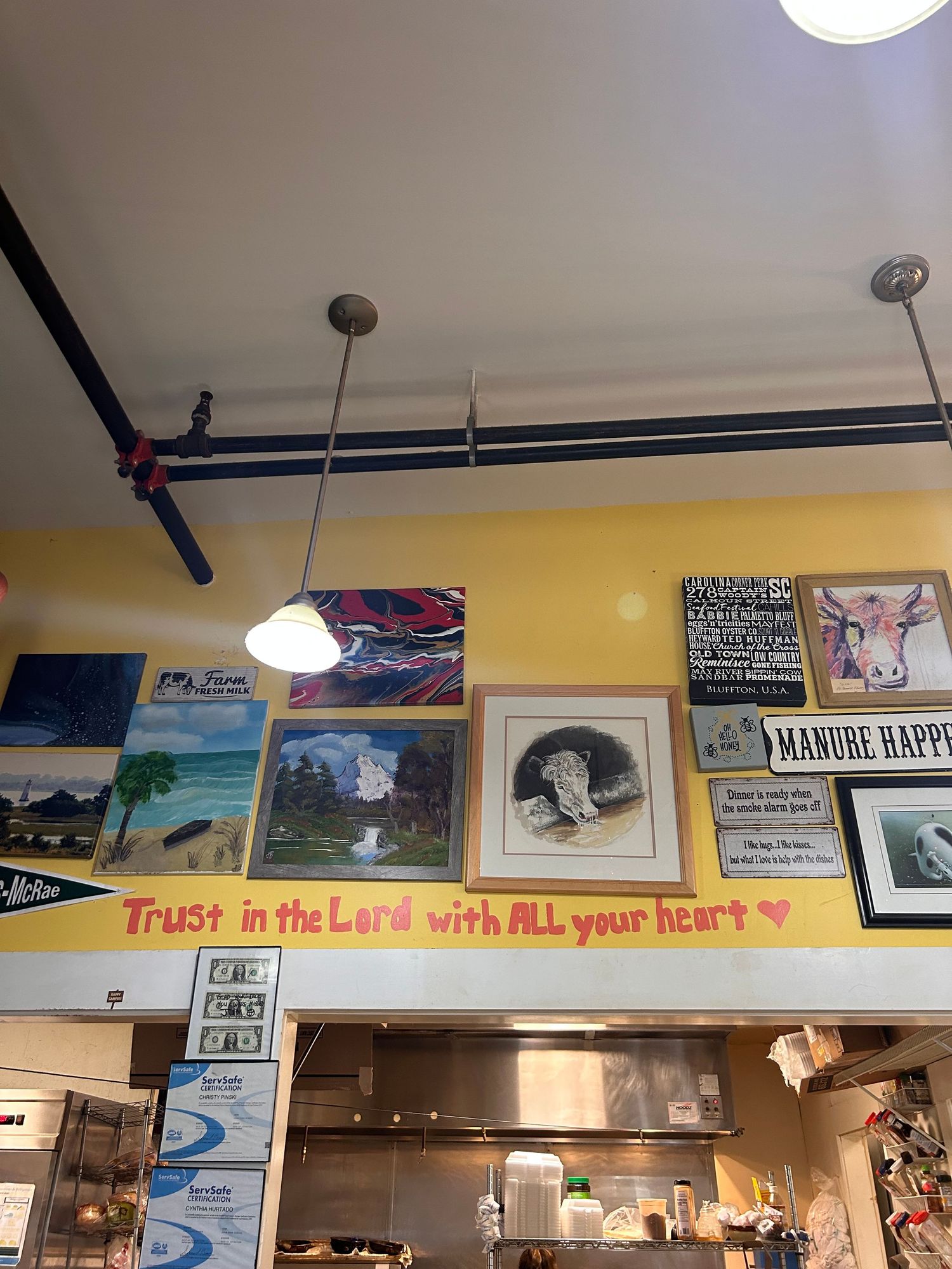 Yellow-walled cafe interior with framed artwork and photos displayed along wall under pendant lights and exposed ceiling pipes.