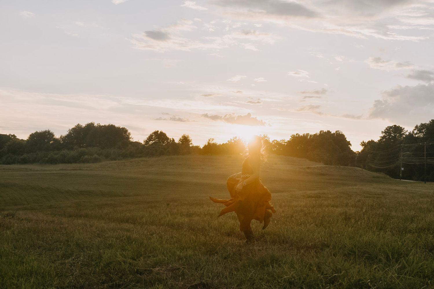 Silhouette of person sitting in field against dramatic sunset with trees in background.
