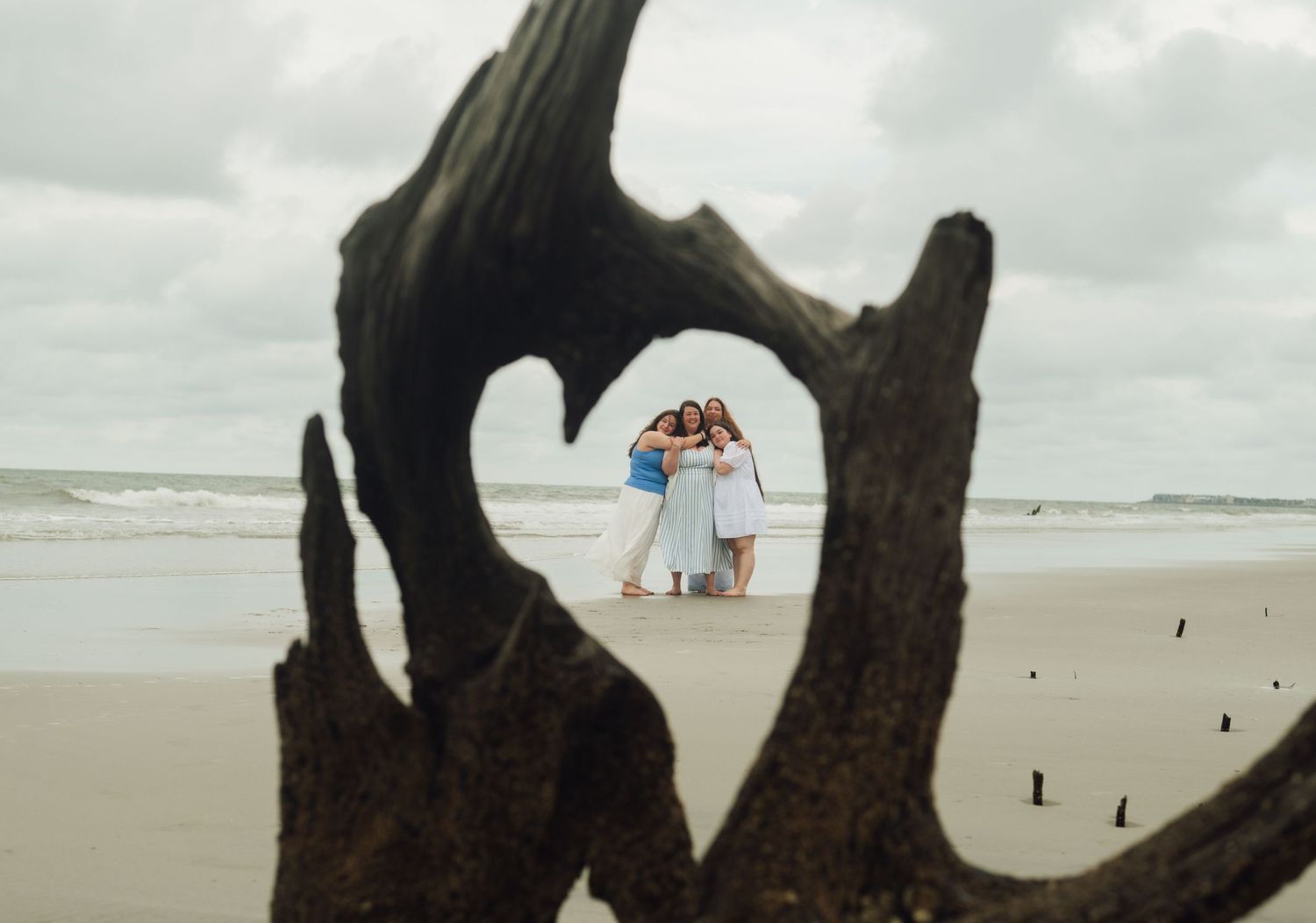 Through a heart-shaped driftwood frame on a beach, a couple stands together under an umbrella by the ocean.