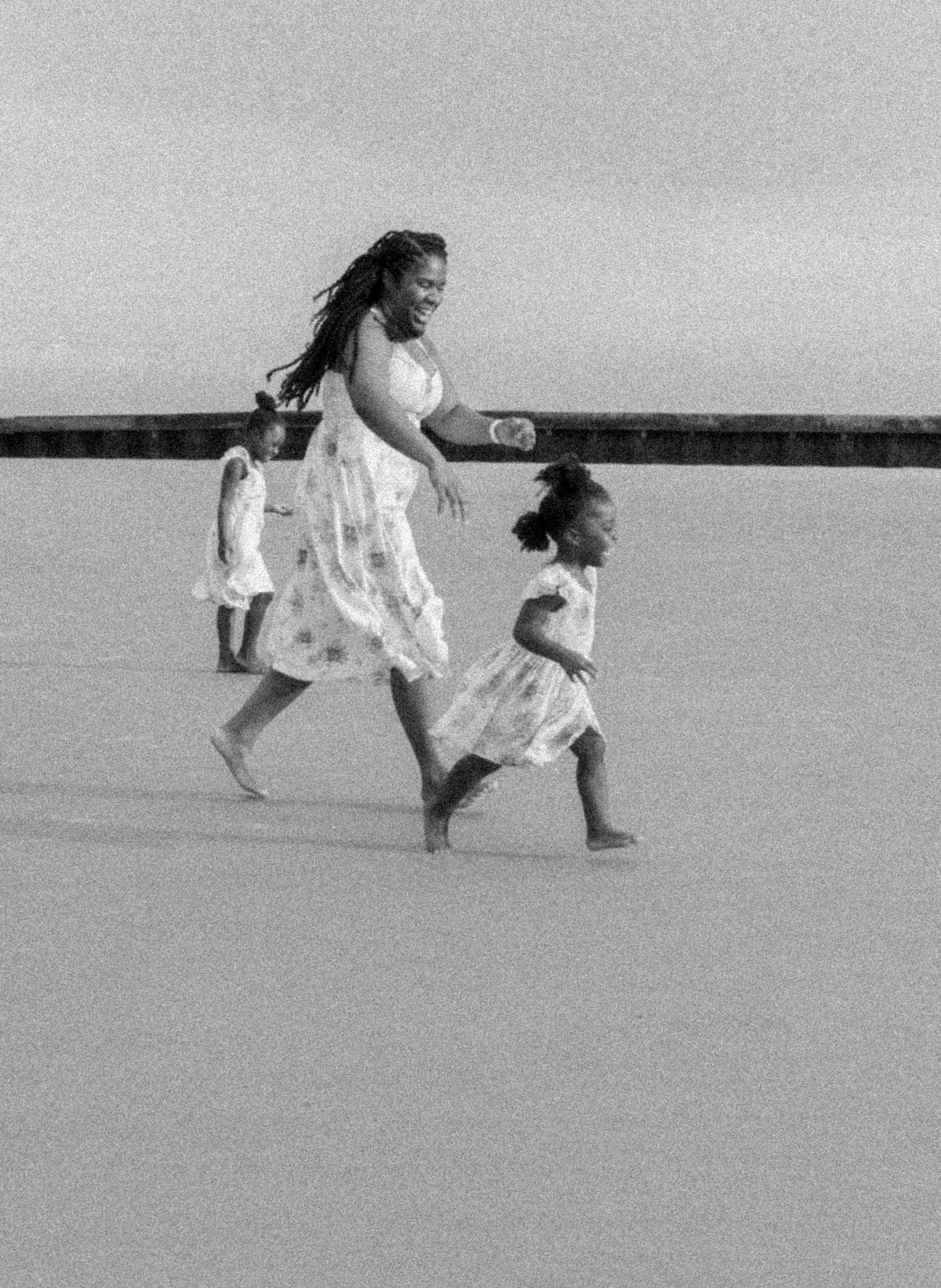 A black and white photograph shows two people in flowing white dresses running joyfully along a beach.