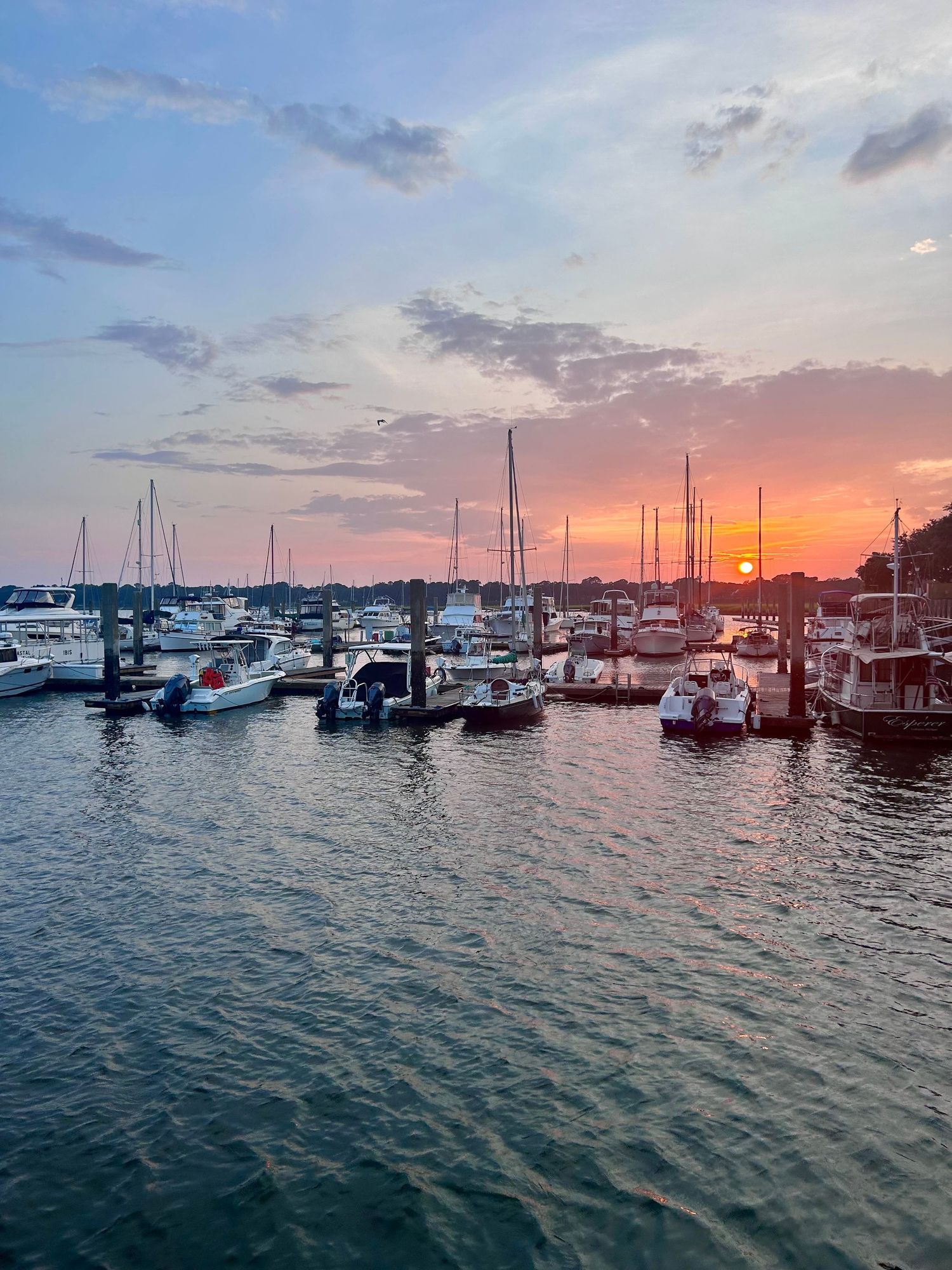 Boats and yachts docked at a marina during a vibrant sunset with orange and pink clouds reflecting on calm waters.
