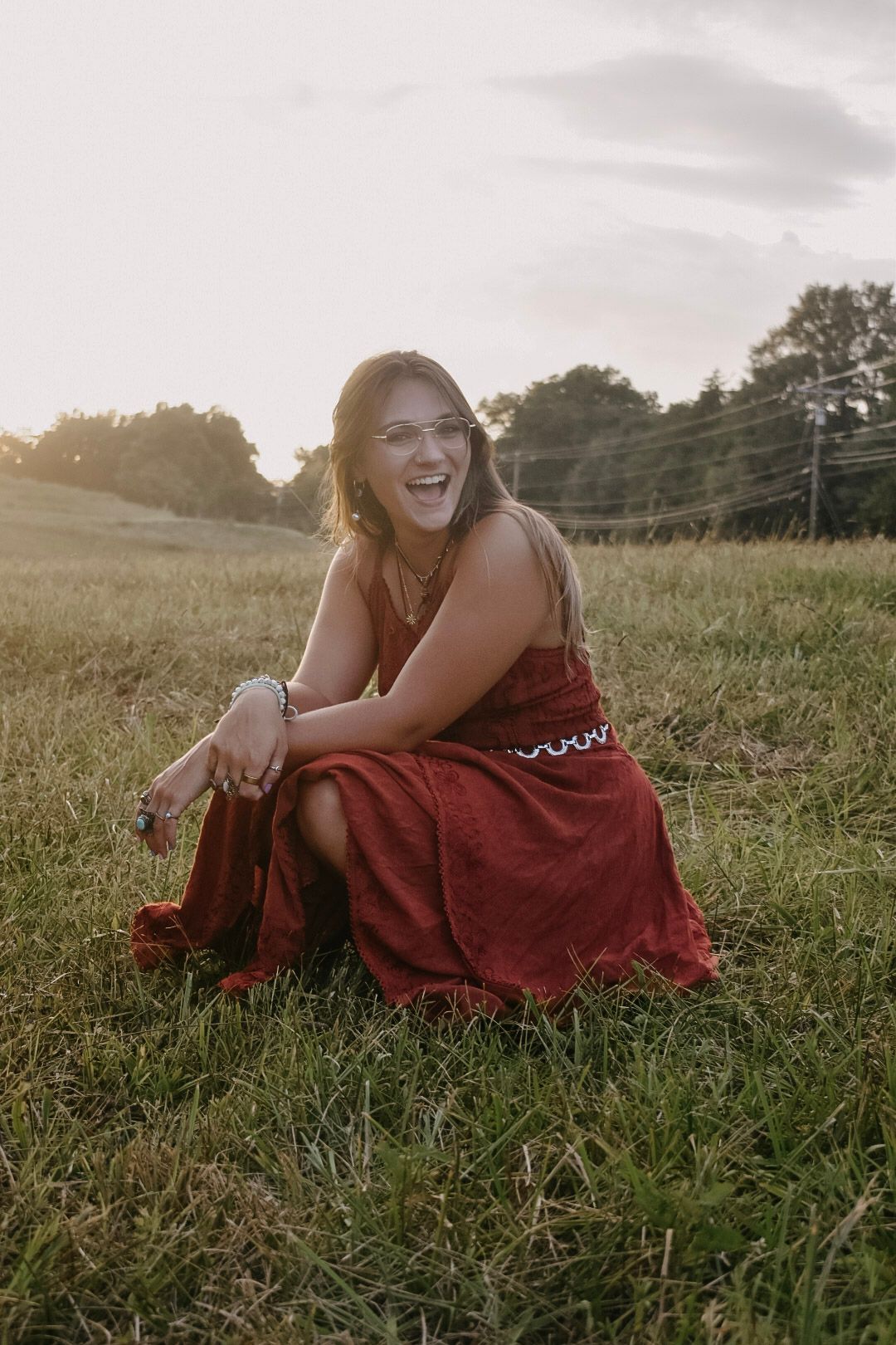A person in a flowy red dress sits in a golden sunlit field during sunset, laughing with joy.