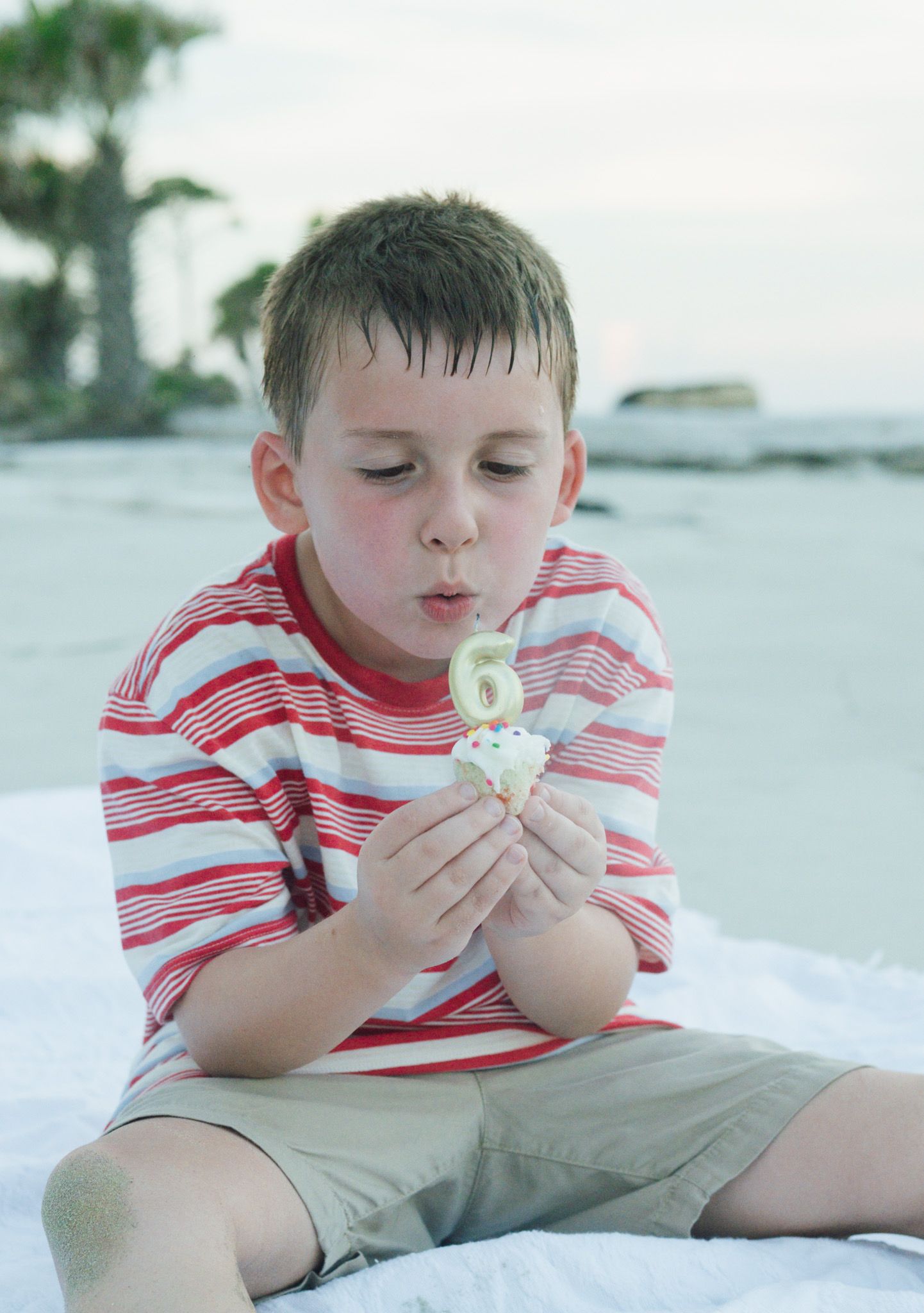 Young child in red and white striped shirt sitting on beach blowing a dandelion flower on sunny day.