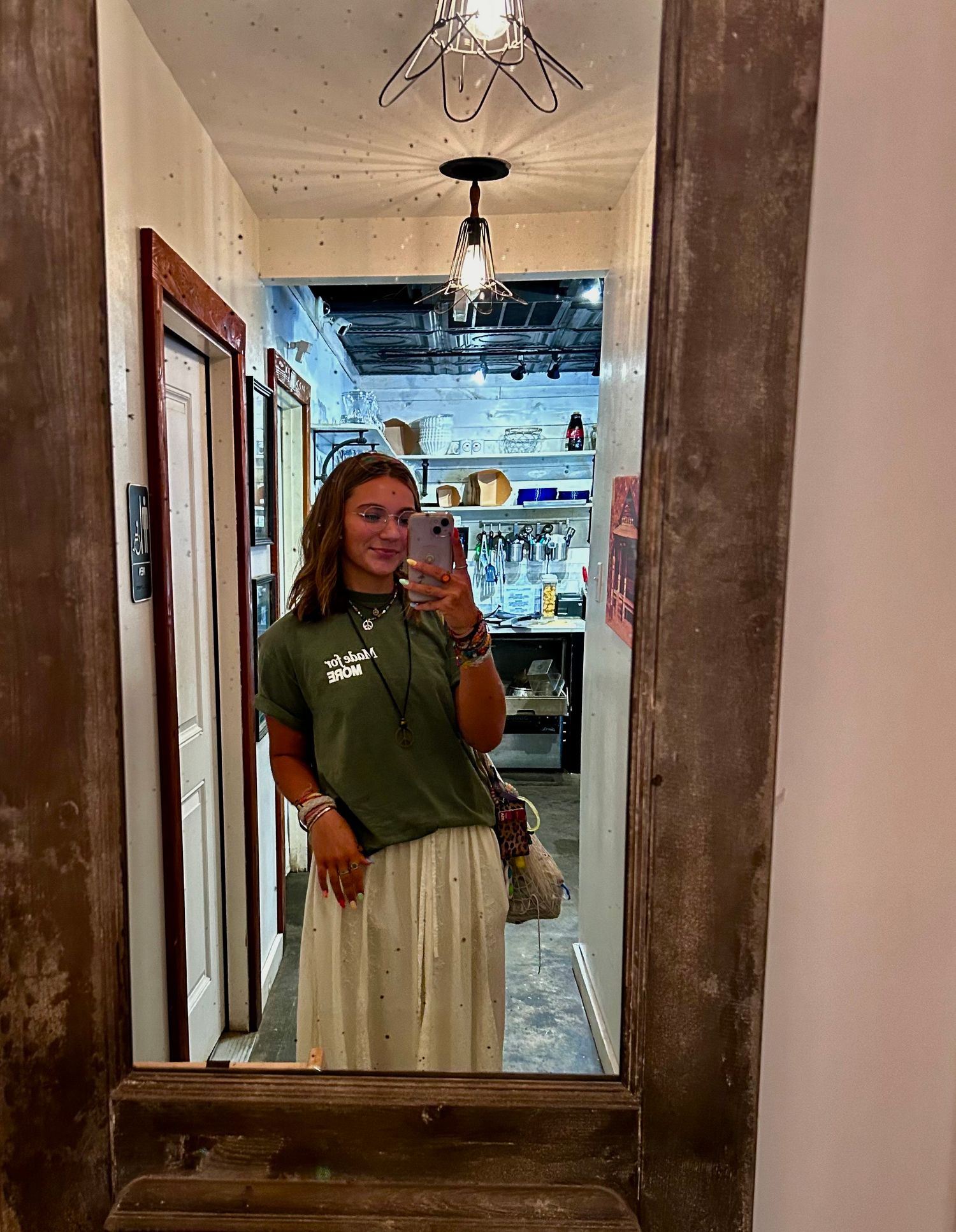 Person wearing olive green t-shirt and light colored pants poses in front of industrial-style mirror in hallway.