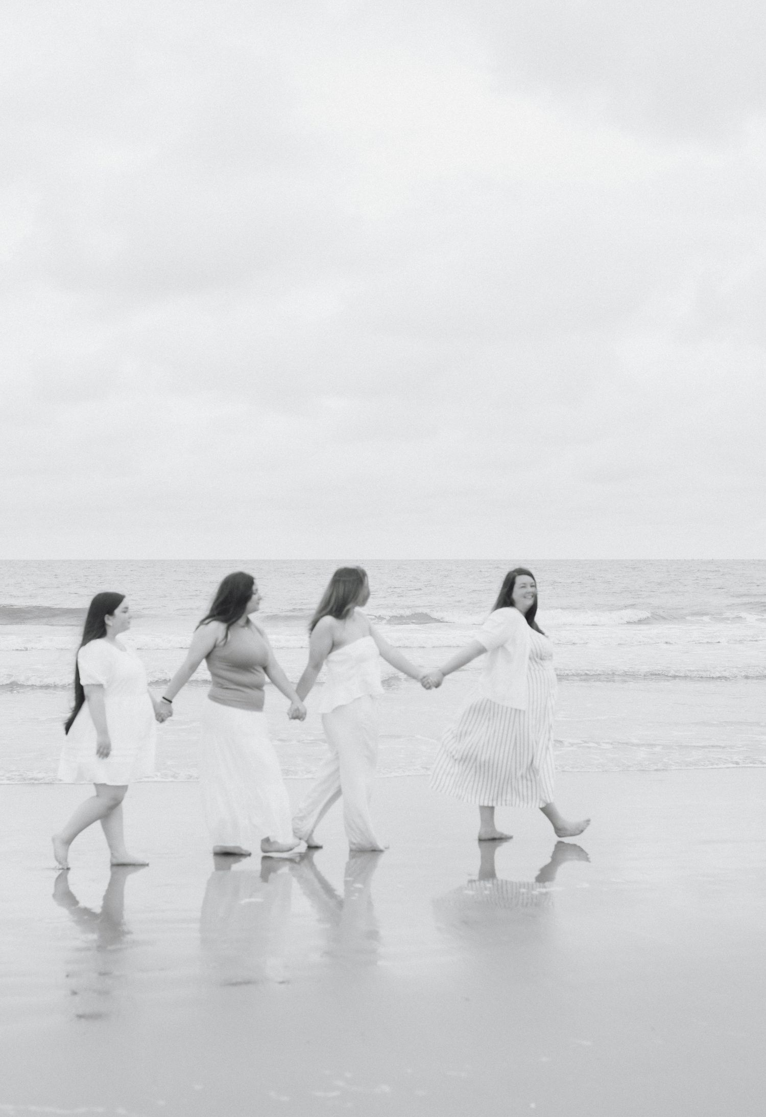 A group in white outfits walks along a misty beach holding hands in a joyful line formation.