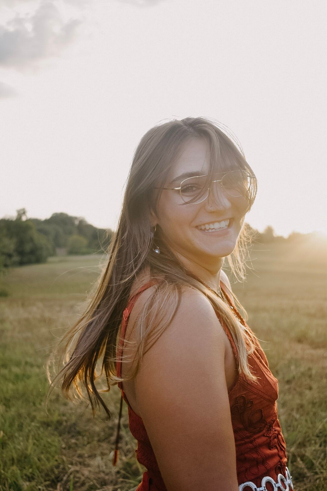 A figure in a red dress smiles warmly in a grassy field during golden hour sunset.