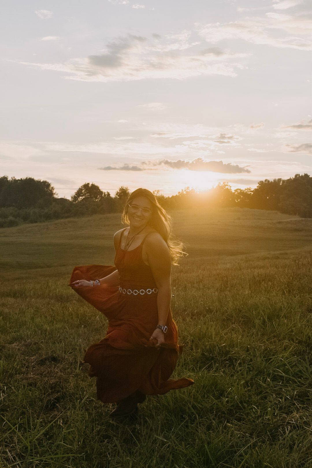 A silhouetted figure sits in a grassy field at golden hour as the sun sets behind trees on the horizon.