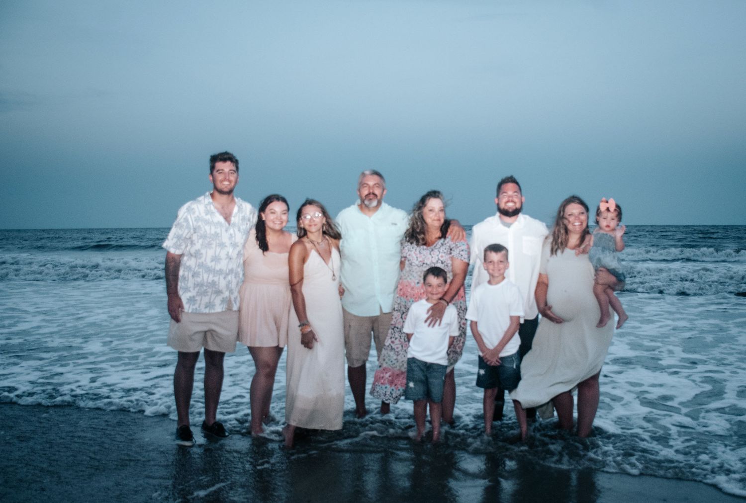 A group of people in light colored clothing stand together at the edge of the ocean during blue hour.