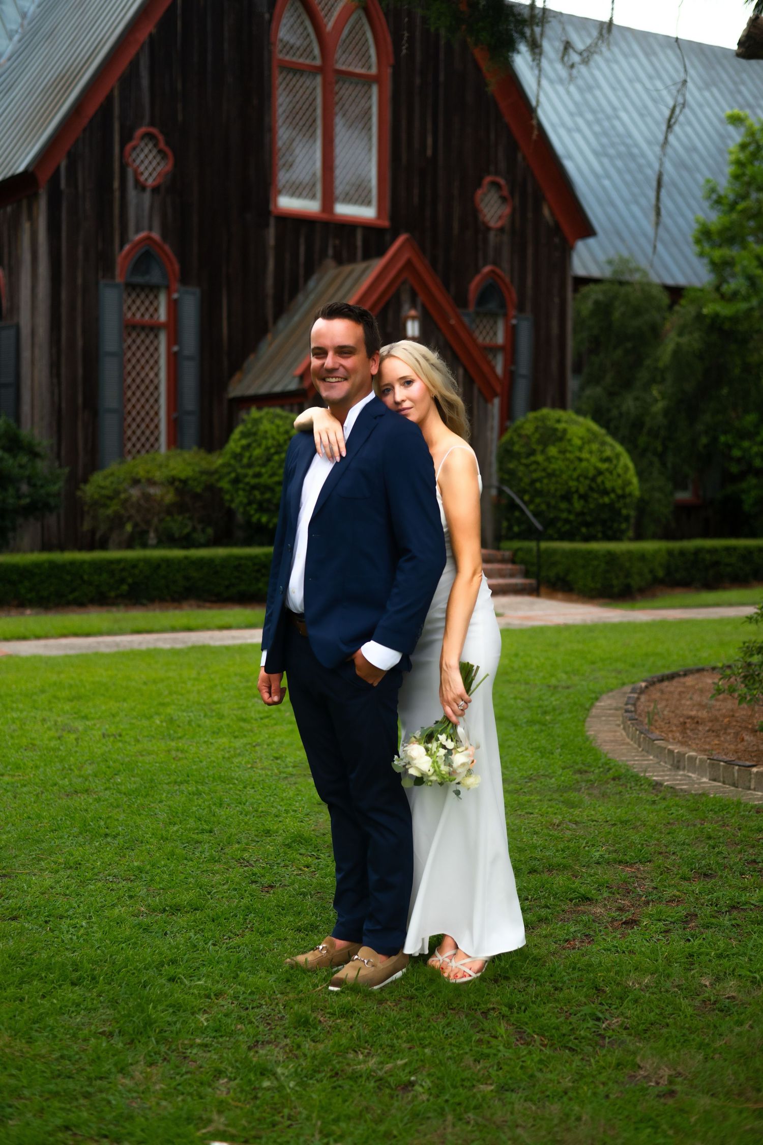 A wedding couple poses on a lawn in front of a charming gothic-style church with arched windows.