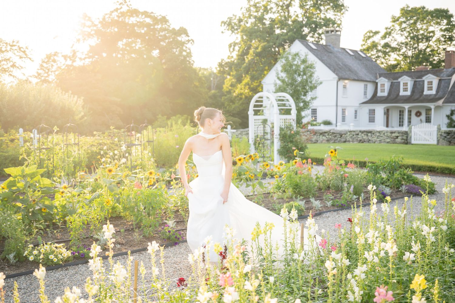 Bride in white gown standing in a sunlit garden with colorful flowers and a white farmhouse in the background.