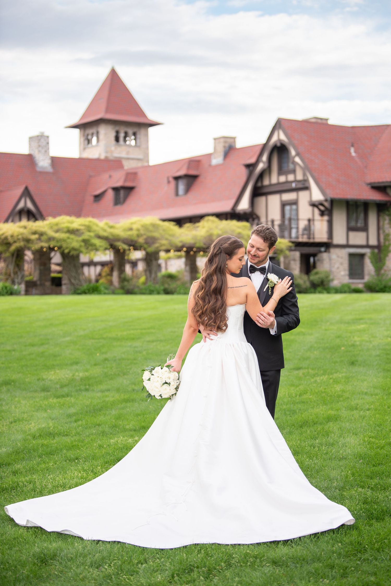 Bride in white gown and groom in black tux embrace on green lawn before Tudor-style wedding venue.