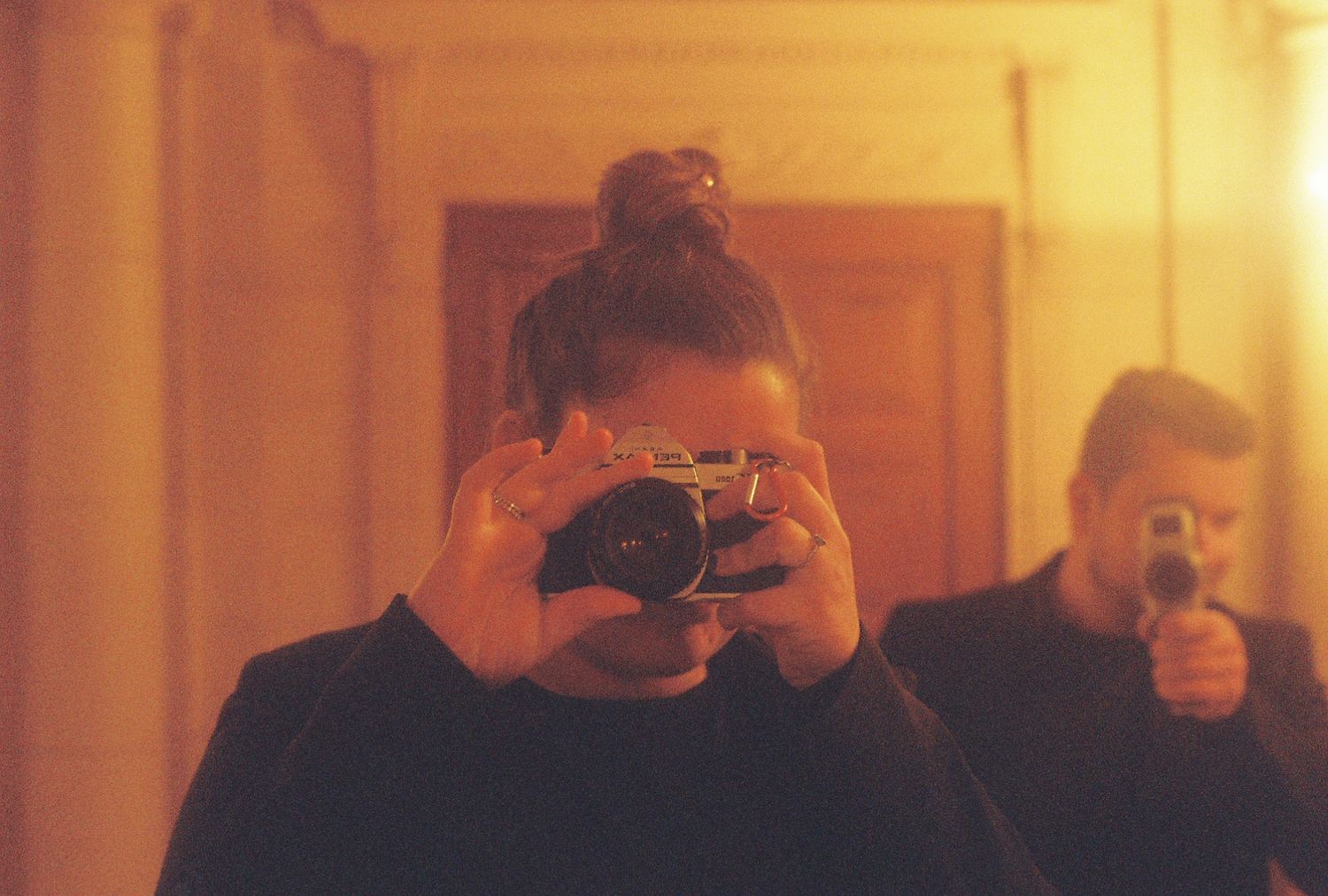 Someone taking a photo with a vintage camera while another person stands in the background in warm lighting.