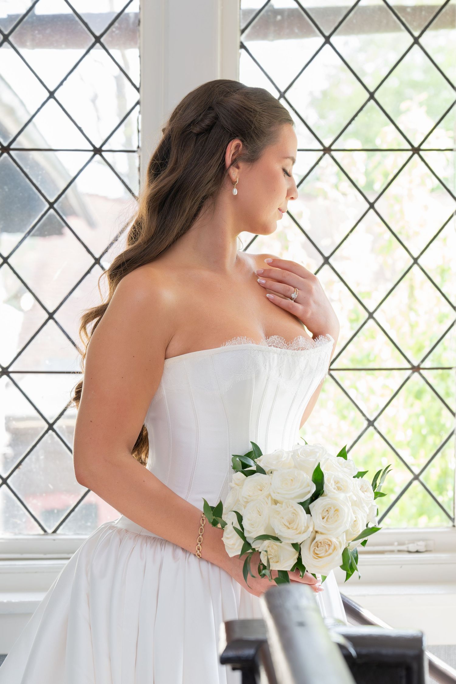 Elegant bride in strapless white gown holding bouquet of white roses, standing by diamond-patterned window.