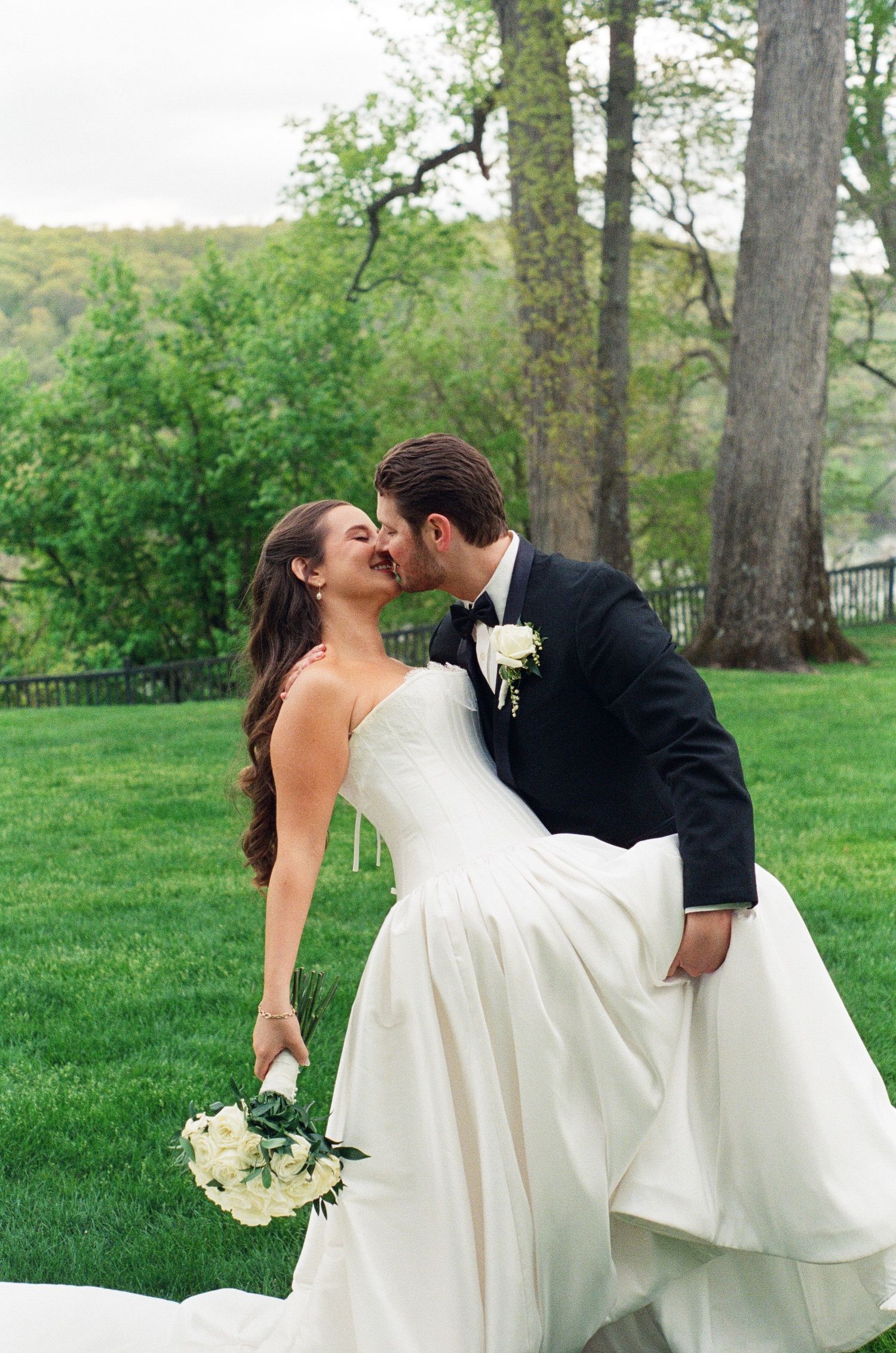 A groom in a black tuxedo dips and kisses his bride in a strapless white gown holding a white bouquet outdoors.
