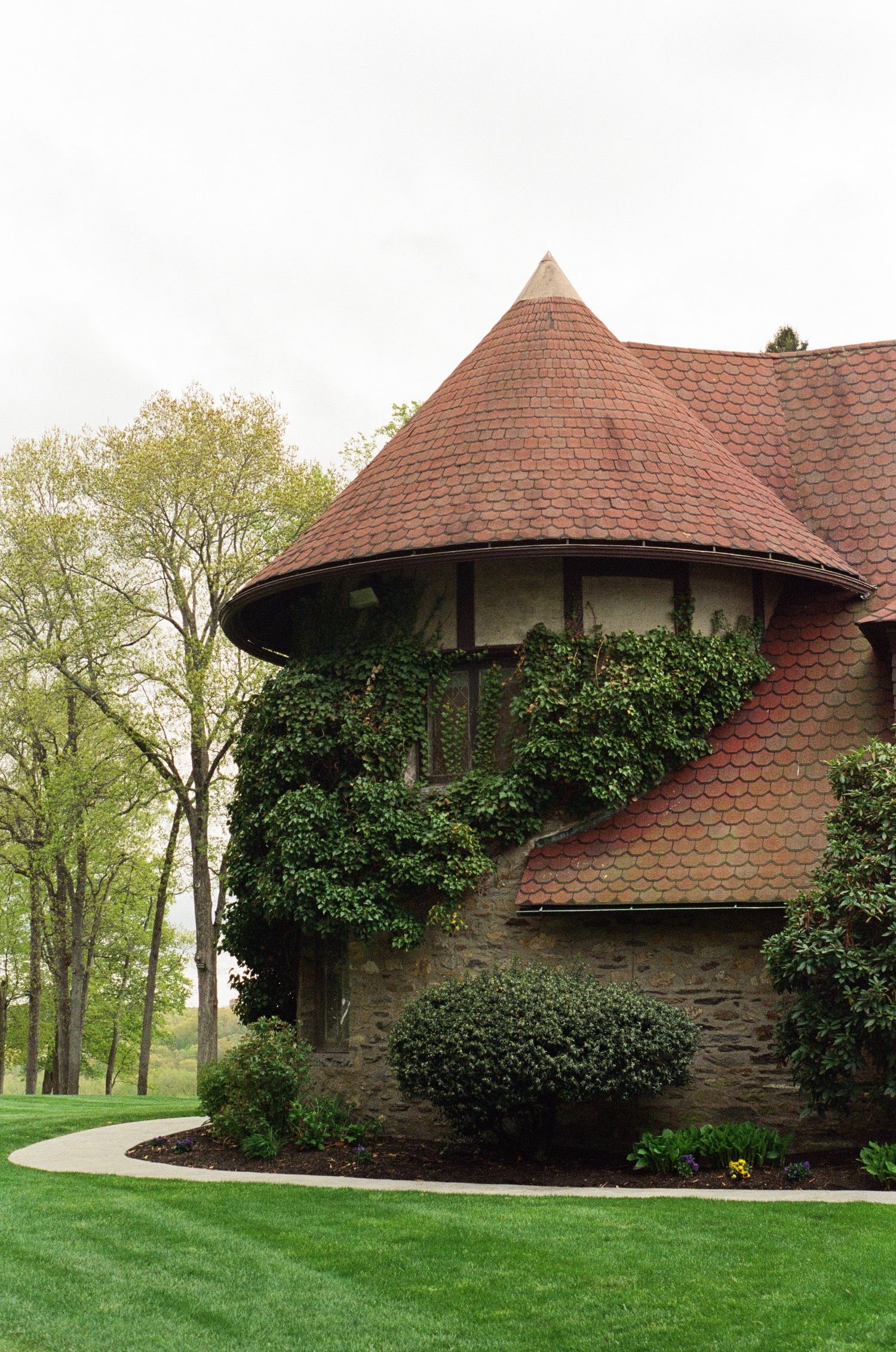 Stone turret building with red conical roof, ivy-covered walls, manicured shrubs, and lush green lawn surrounded by trees.