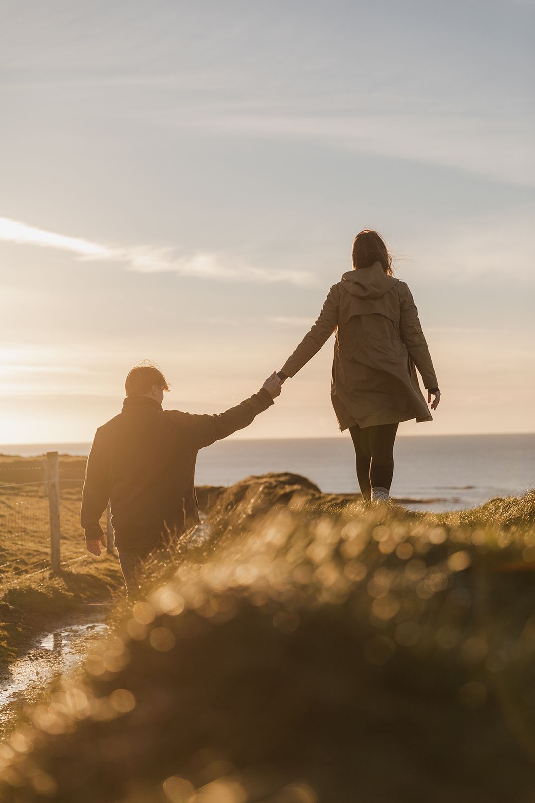 couple are walking Doolin cliff walk towards Cliffs of Moher at sunset during their cinematic, storytelling photo session in Ireland