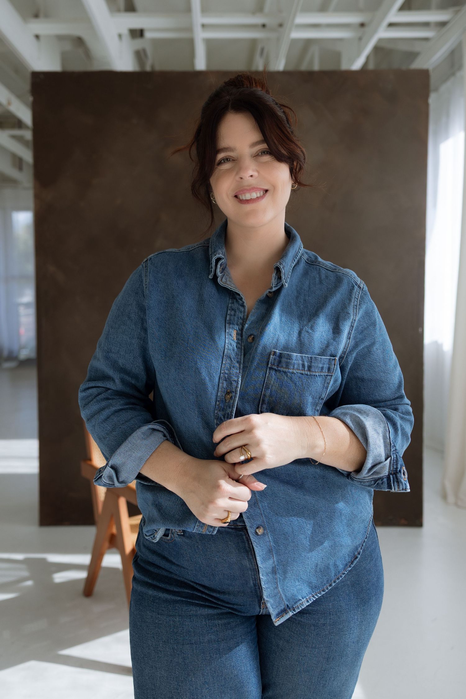Smiling woman in blue denim shirt and jeans posing in a bright modern studio with brown backdrop.