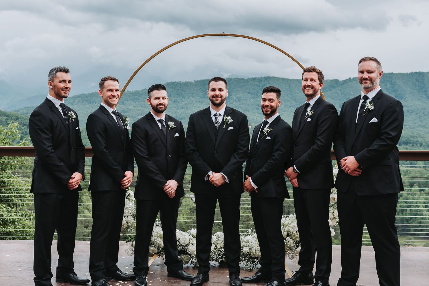 A group of groomsmen in black suits and ties pose together on a scenic mountain overlook with misty hills behind them.