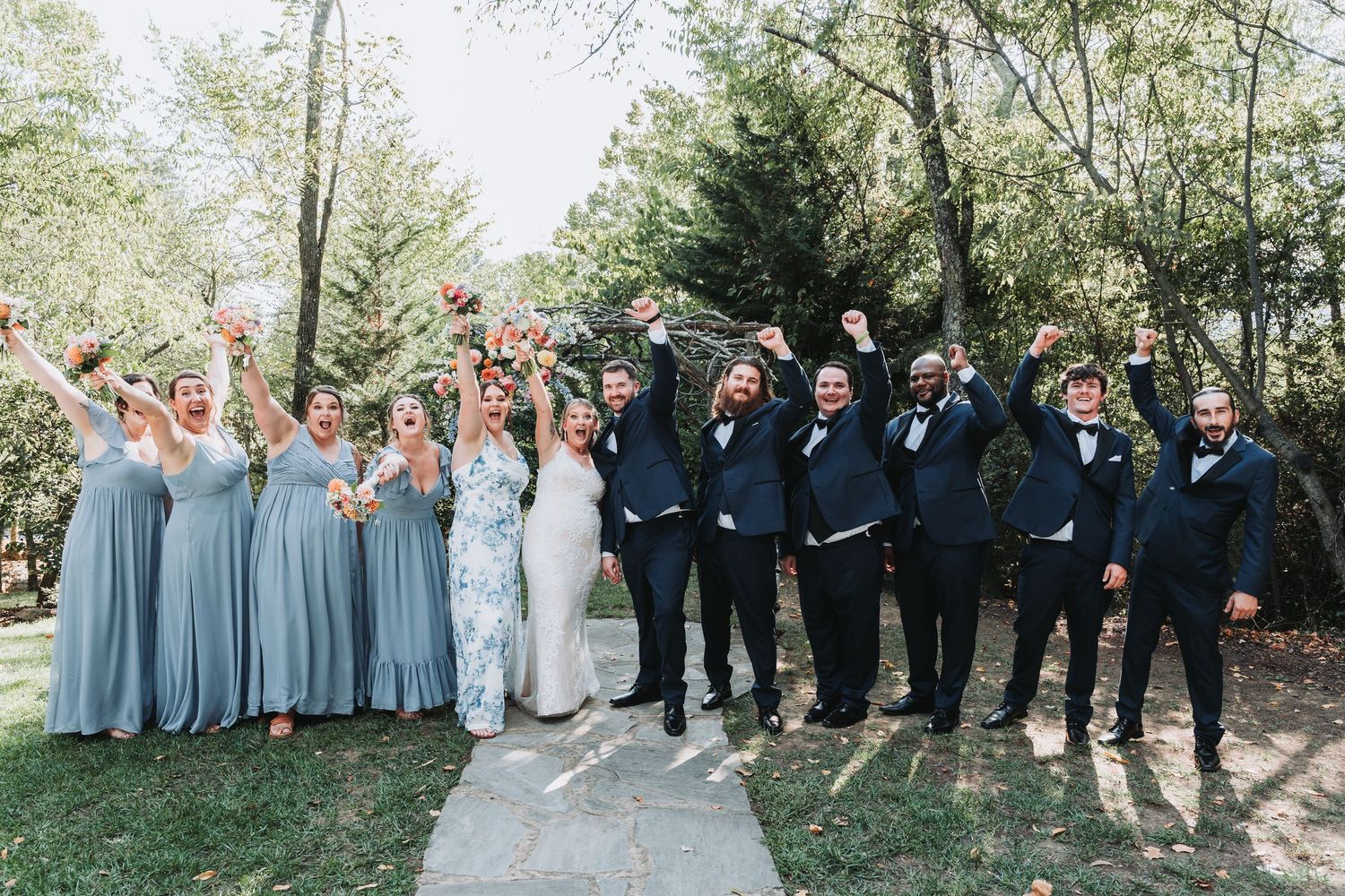 Wedding party stands in line outdoors with bridesmaids in blue dresses and groomsmen in dark suits and bow ties.