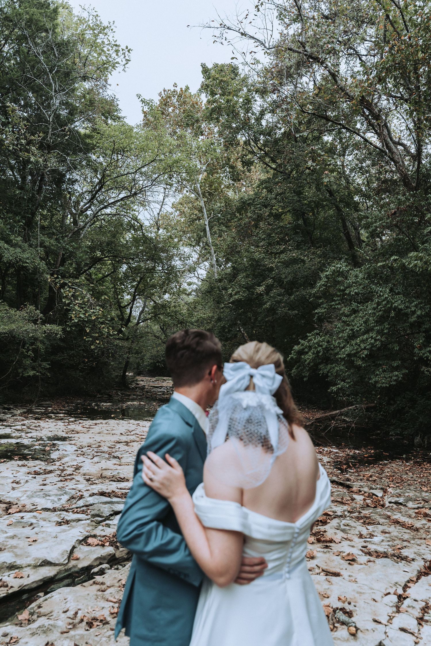 A couple in wedding attire embrace while standing near a creek in a wooded forest setting.