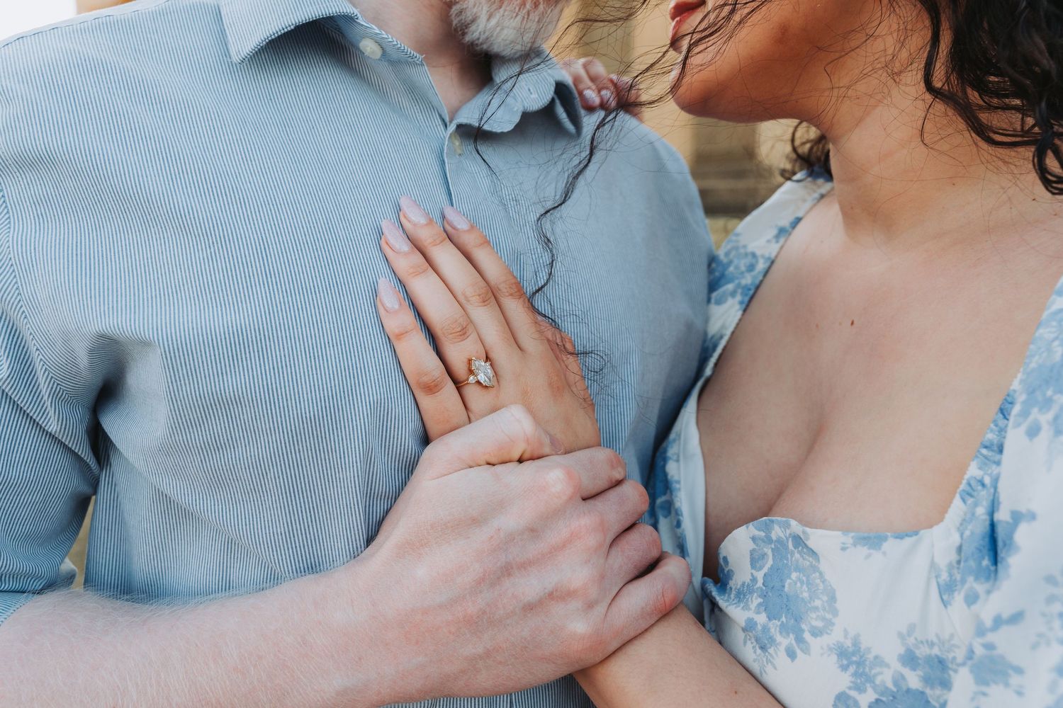 Couple embracing, woman wearing a stunning diamond engagement ring, dressed in blue floral outfit.