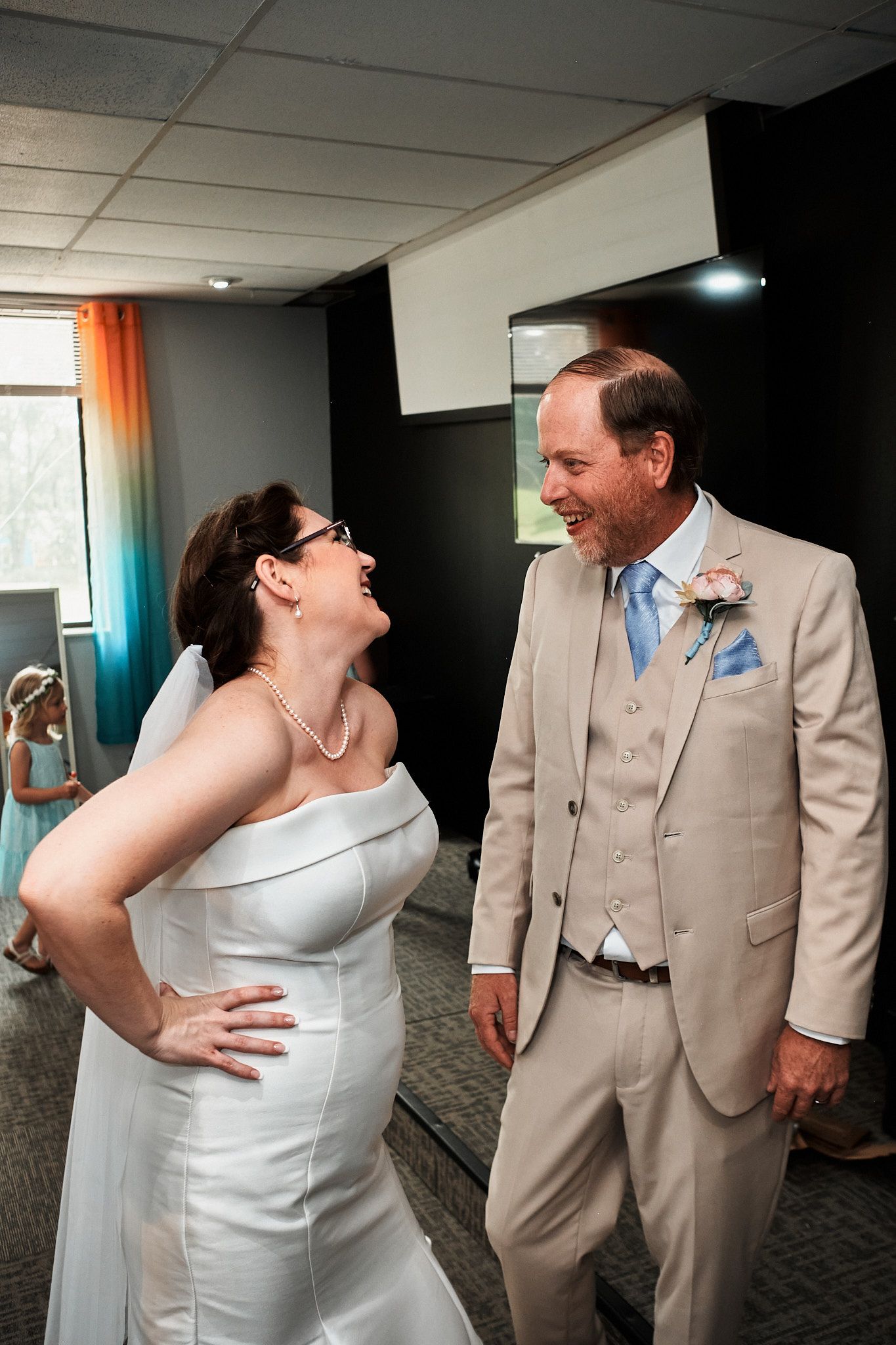 A couple shares a joyful laugh while wearing wedding attire, with the bride in white and groom in tan suit.