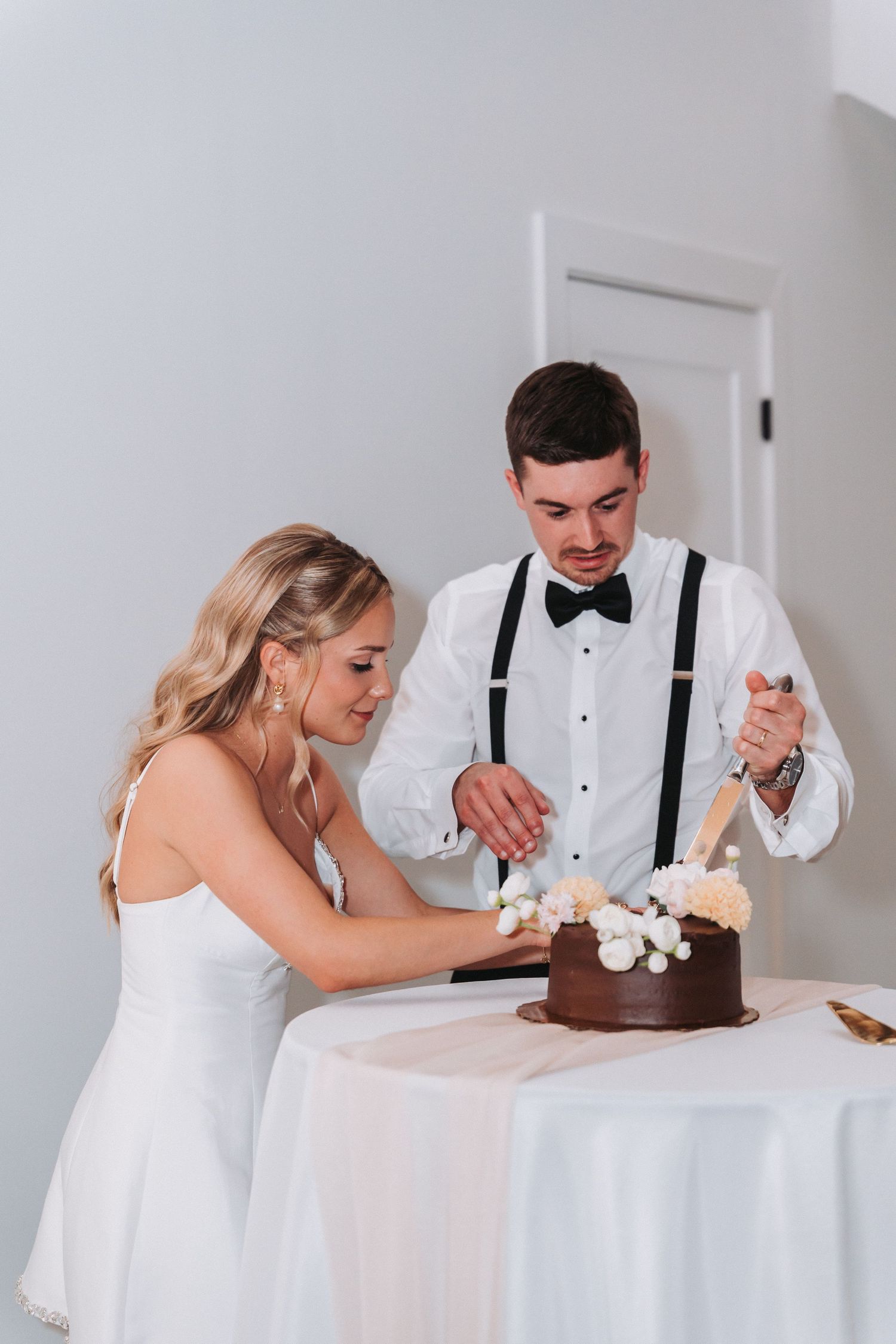Couple in wedding attire cuts into a chocolate cake together at an elegant white table setting during their reception.