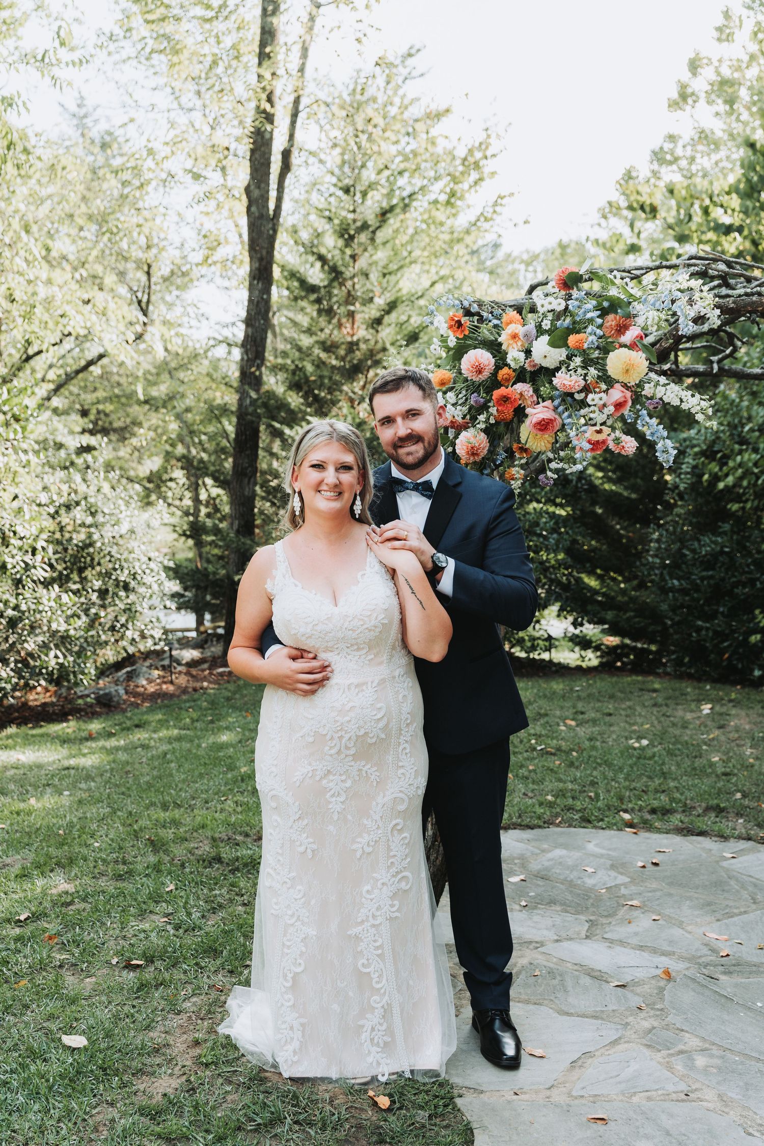 Couple poses together under stunning floral arch with peach roses in sunny outdoor garden.