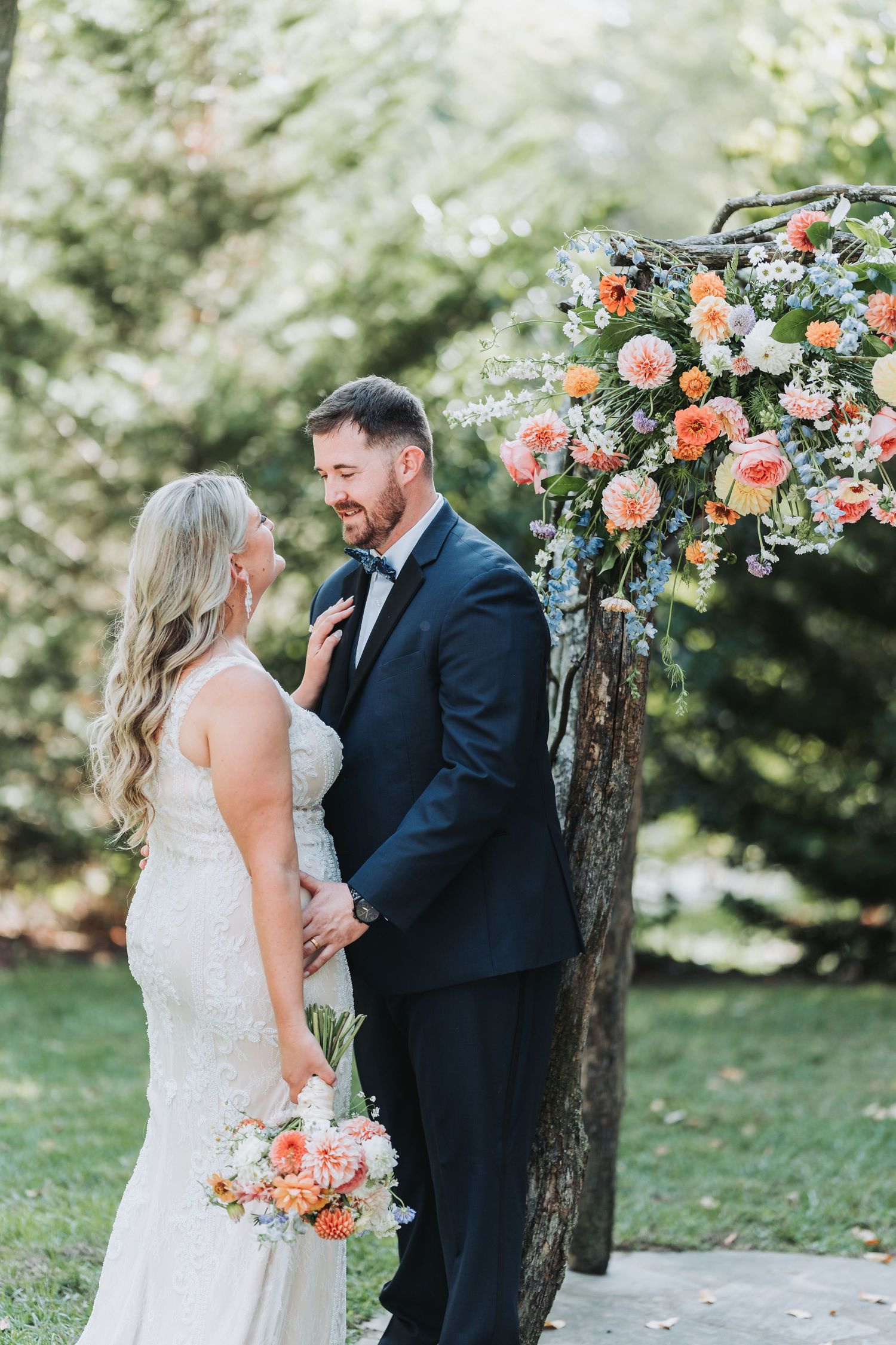 Couple shares romantic moment under beautiful floral arch with peach and coral blooms in outdoor garden setting.